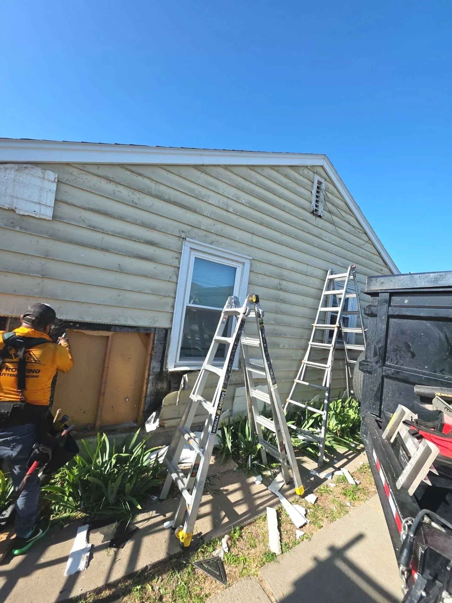 Construction worker repairing house siding, three ladders, and tools on a sidewalk next to a house with yellow siding and a window, under a clear blue sky.