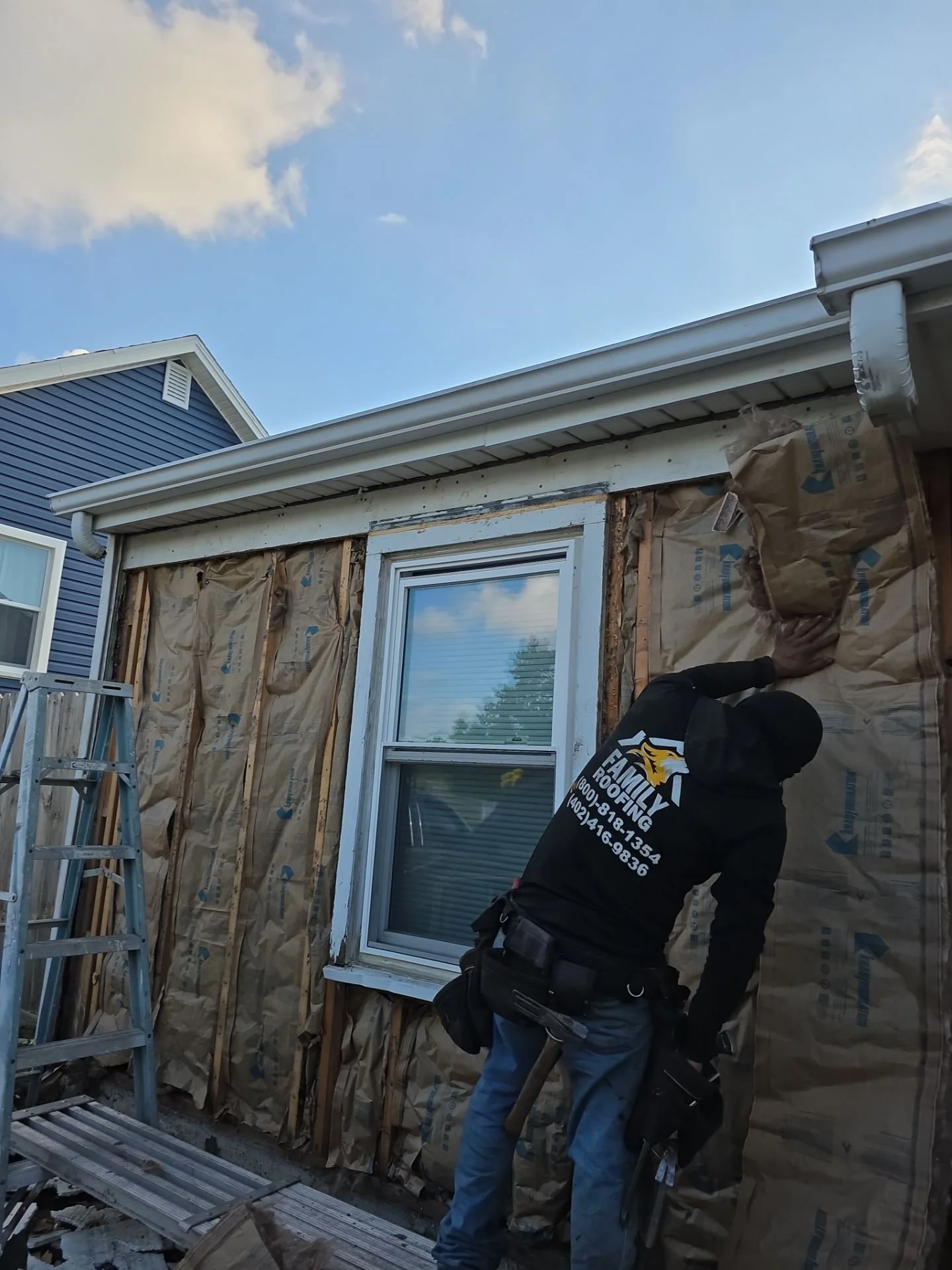 A person working on a house siding renovation, removing old siding and installing new exterior wall insulation, with a ladder nearby and a partly cloudy sky above.