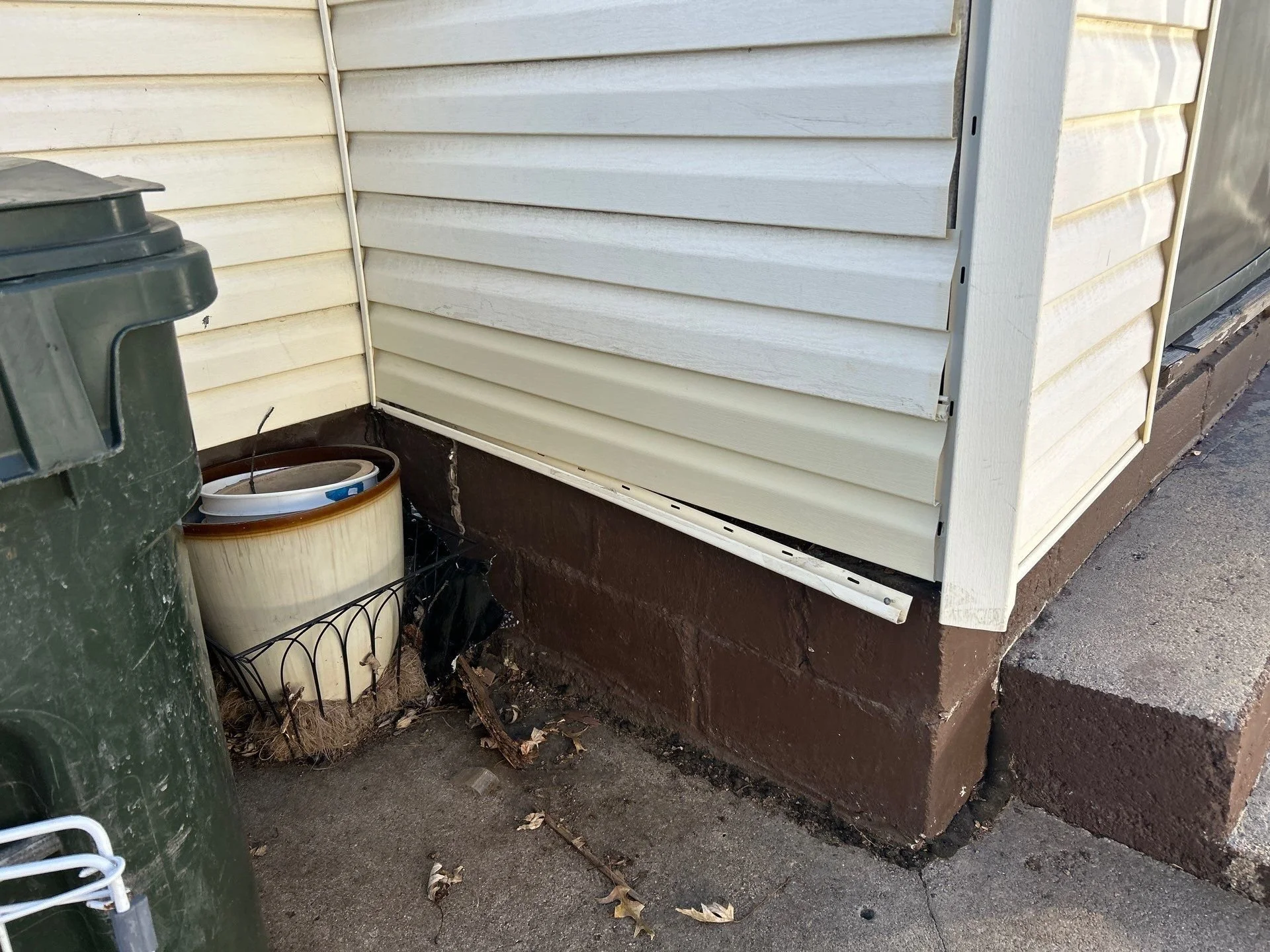 A corner of a house with off-white vinyl siding, a brown concrete foundation, and a rubber gutter. There is a green trash bin and a planter with a white bucket inside, placed on a small wire stand, with some leaves and debris on the ground.
