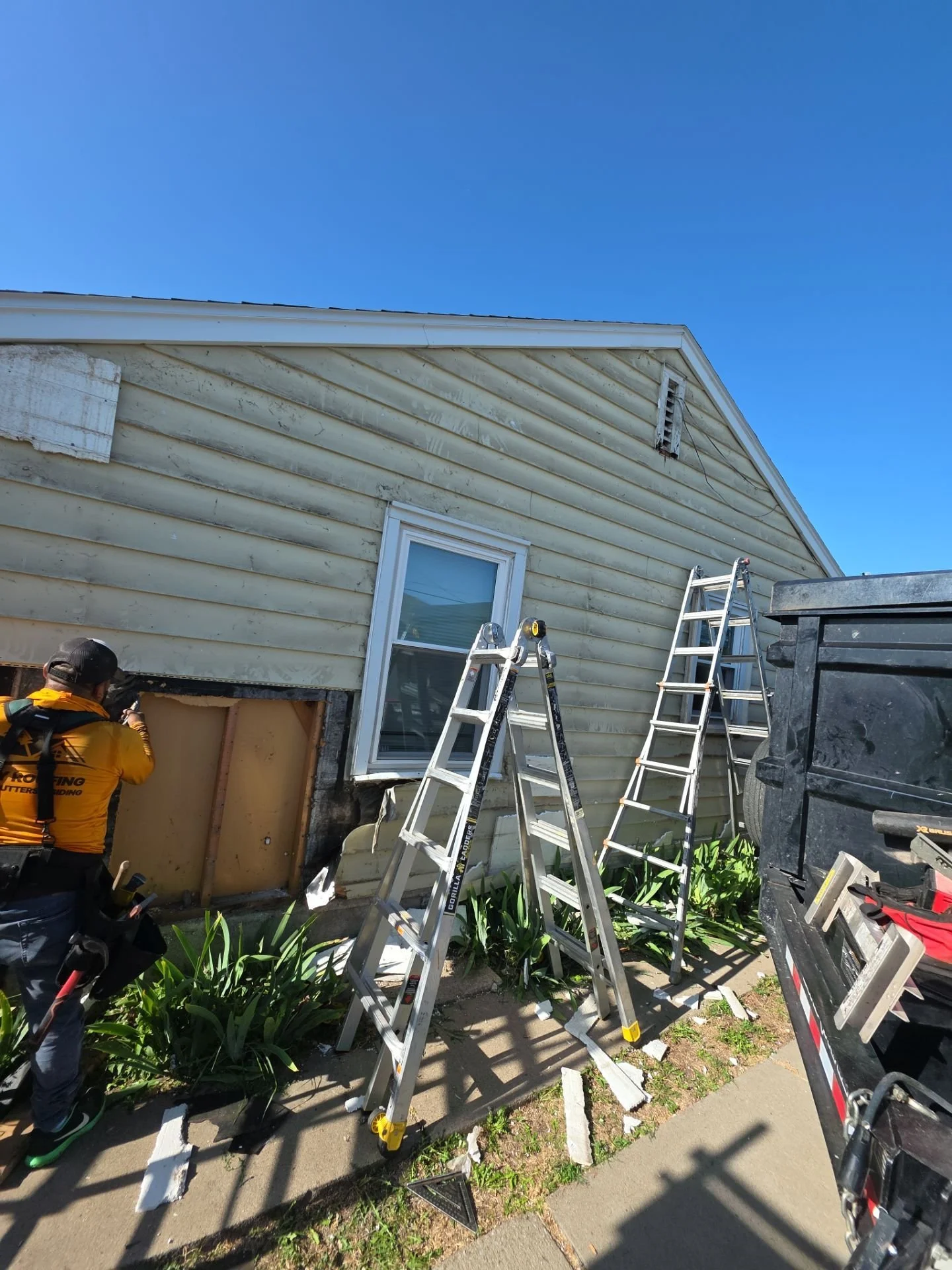 A man working on the exterior of a house with a damaged wall, two ladders, a large black trash bin, some tools, and debris on the sidewalk under a blue sky.