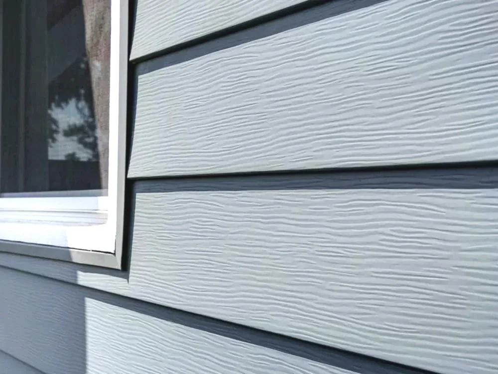 Close-up of house siding in light gray with textured wood grain pattern, adjacent to a white-framed window.