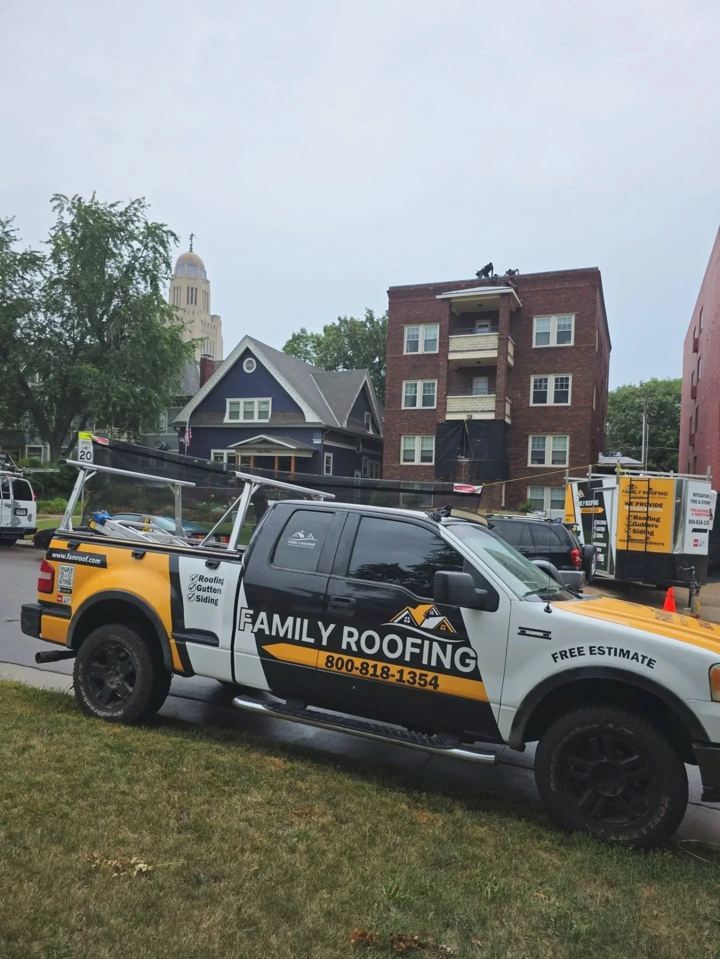 A black and yellow family roofing truck parked on a grassy area in front of a construction site with a three-story brick building and a blue house, with trees and a tall government building in the background.