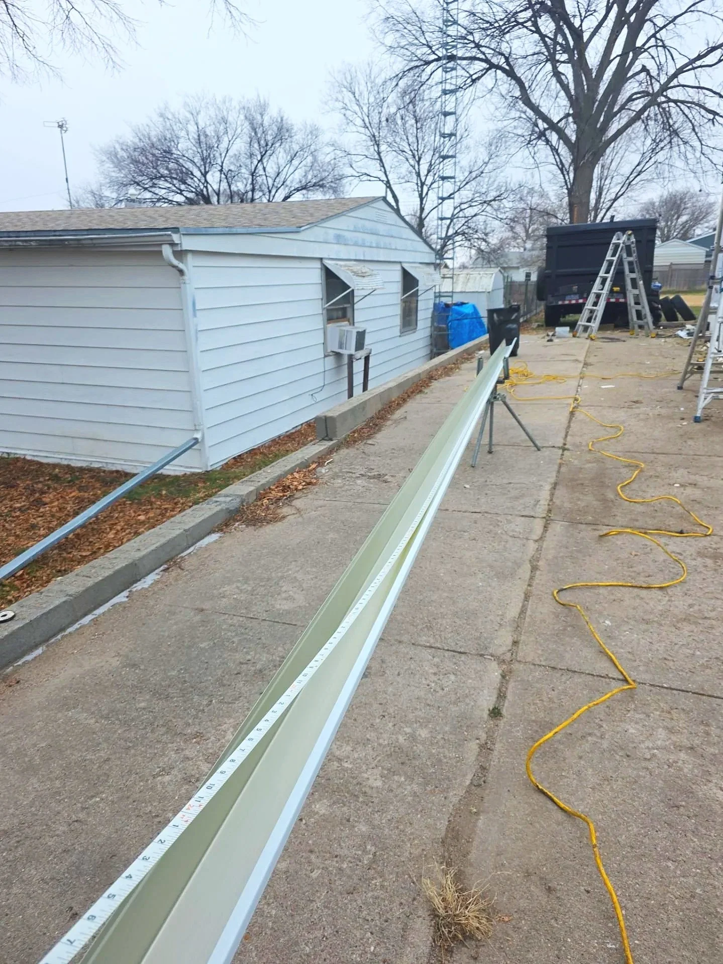 Construction site with a long aluminum beam on a concrete driveway, ladders, a black truck, and trees in the background, during overcast weather.