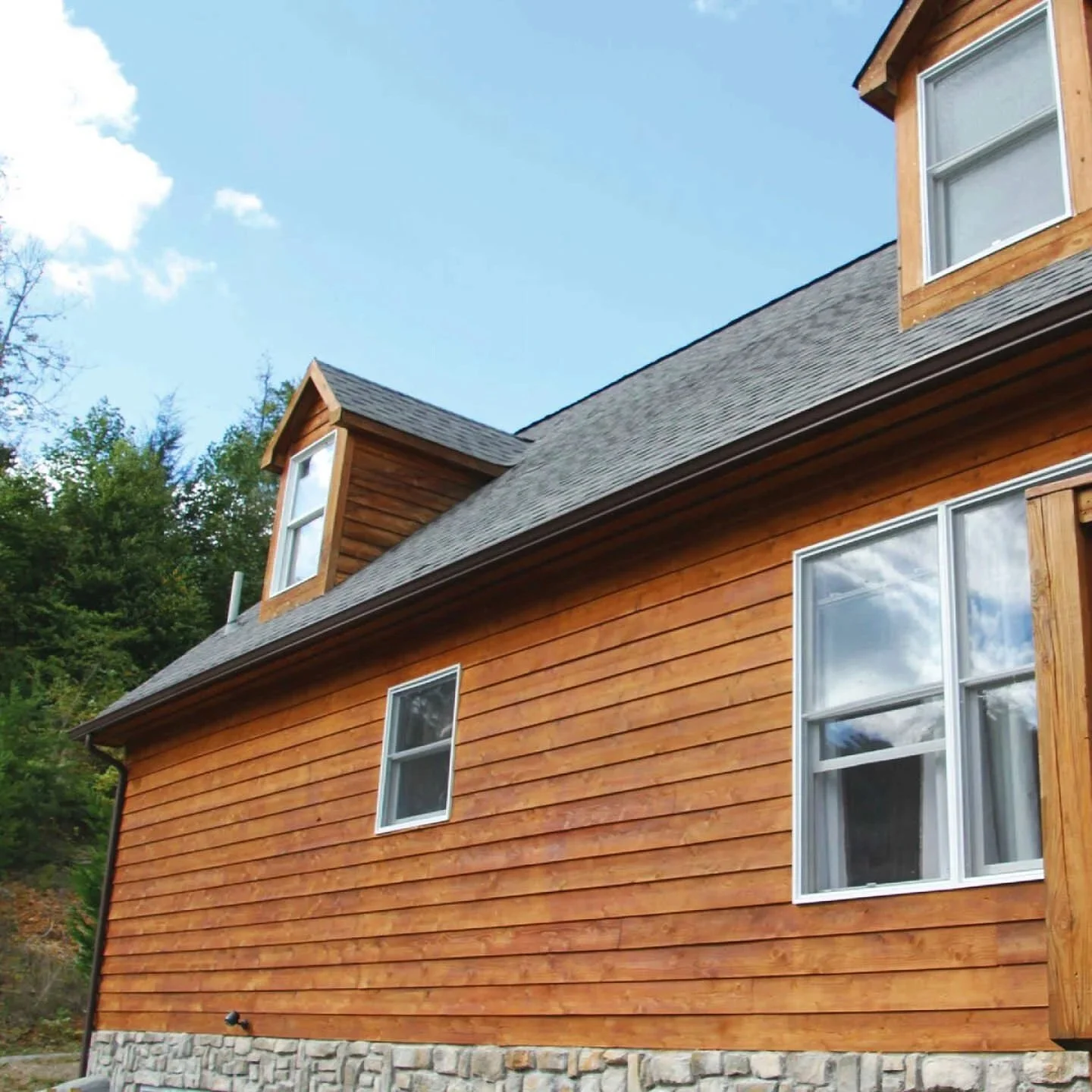 Side view of a wooden house with three windows, a stone foundation, and a shingled roof, surrounded by trees and under a blue sky with clouds.