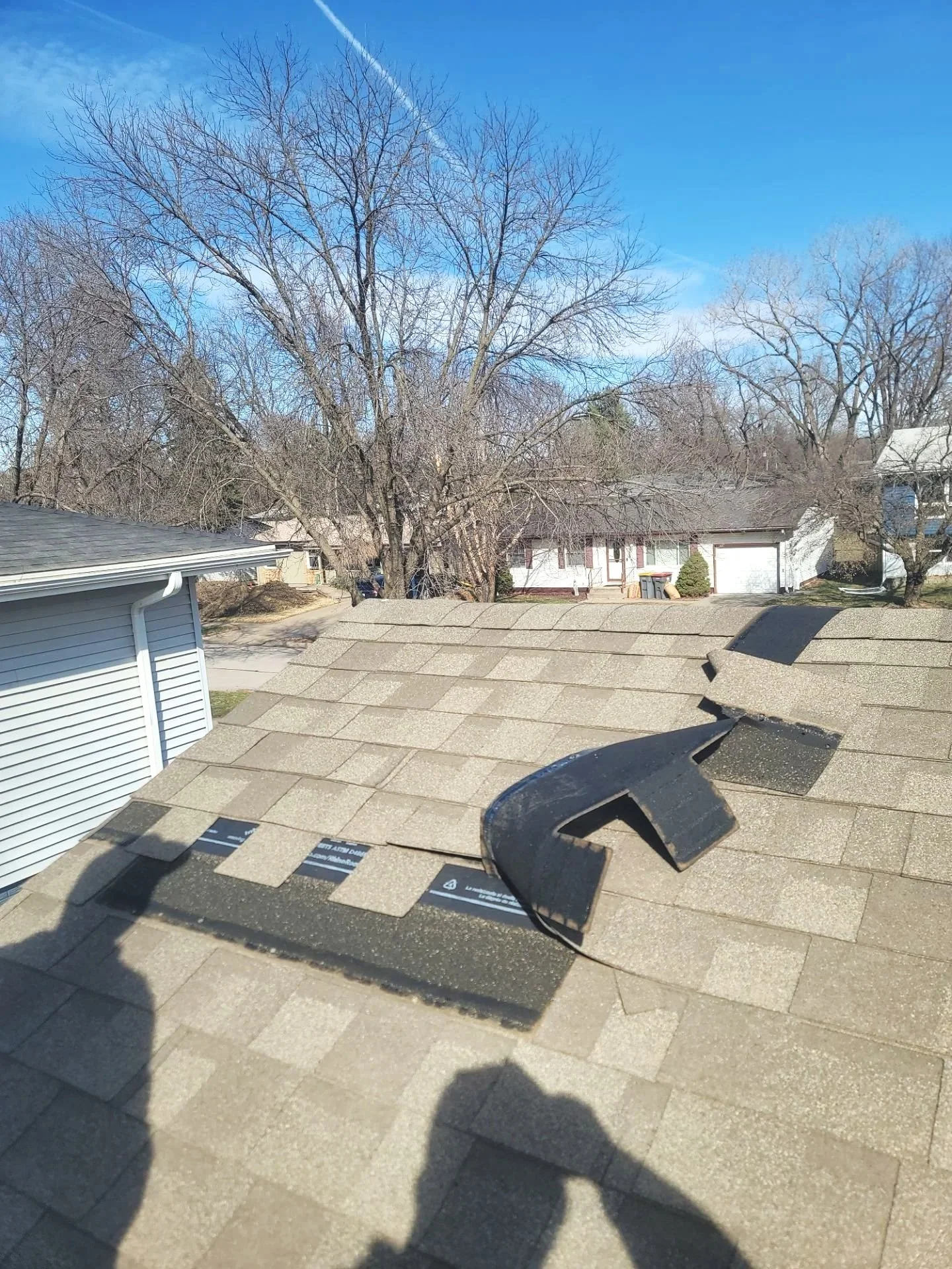 View from a roof showing shingles, a black vent pipe flashing, a clear sky, and neighboring houses with trees.
