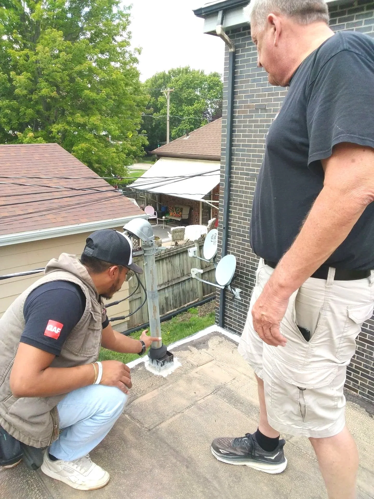 Two men are inspecting a chimney pipe on a roof; one man is kneeling and holding the pipe, while the other man stands nearby, watching.