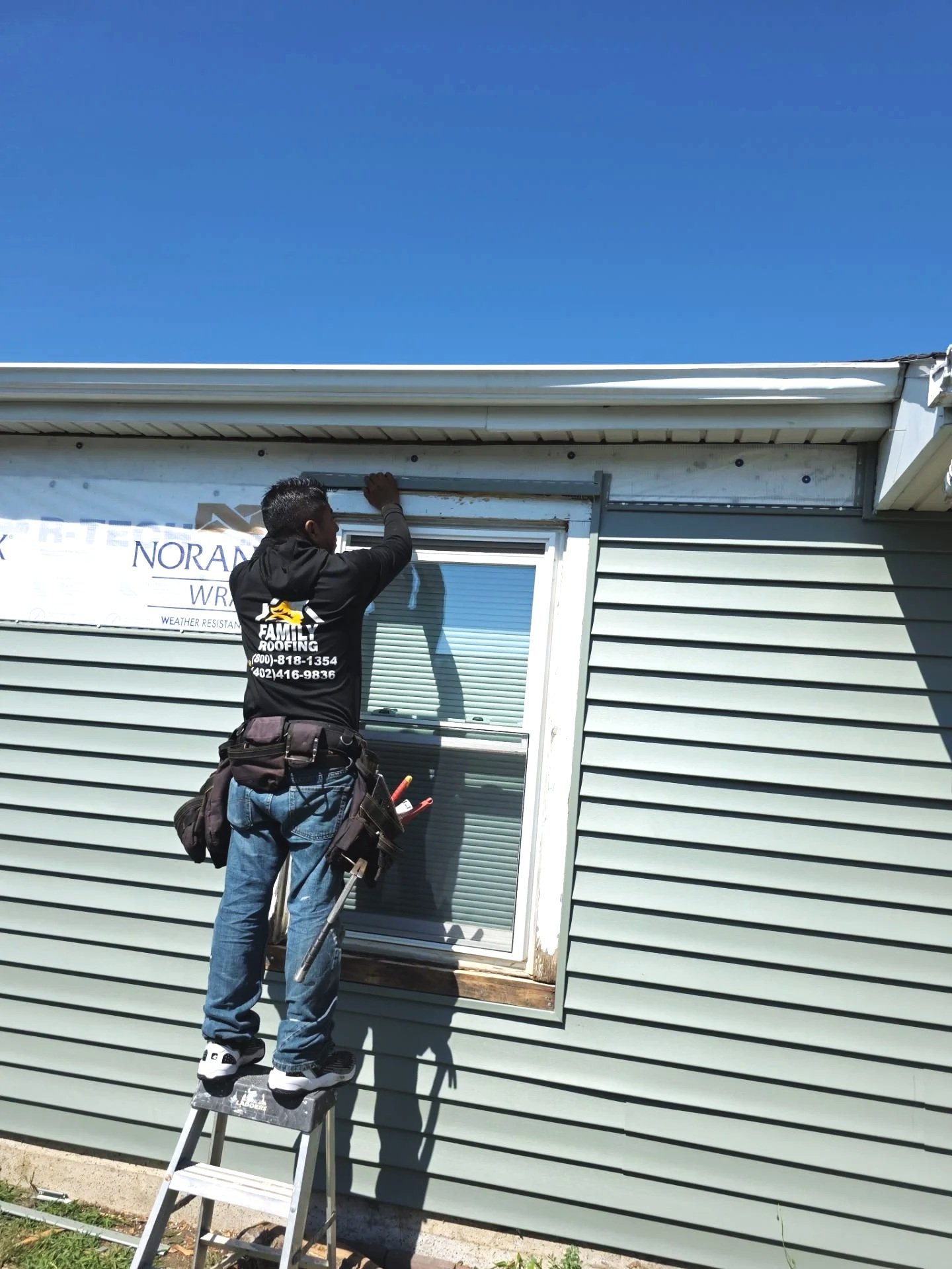 A man standing on a ladder installing or repairing a window on the side of a house under a clear blue sky.