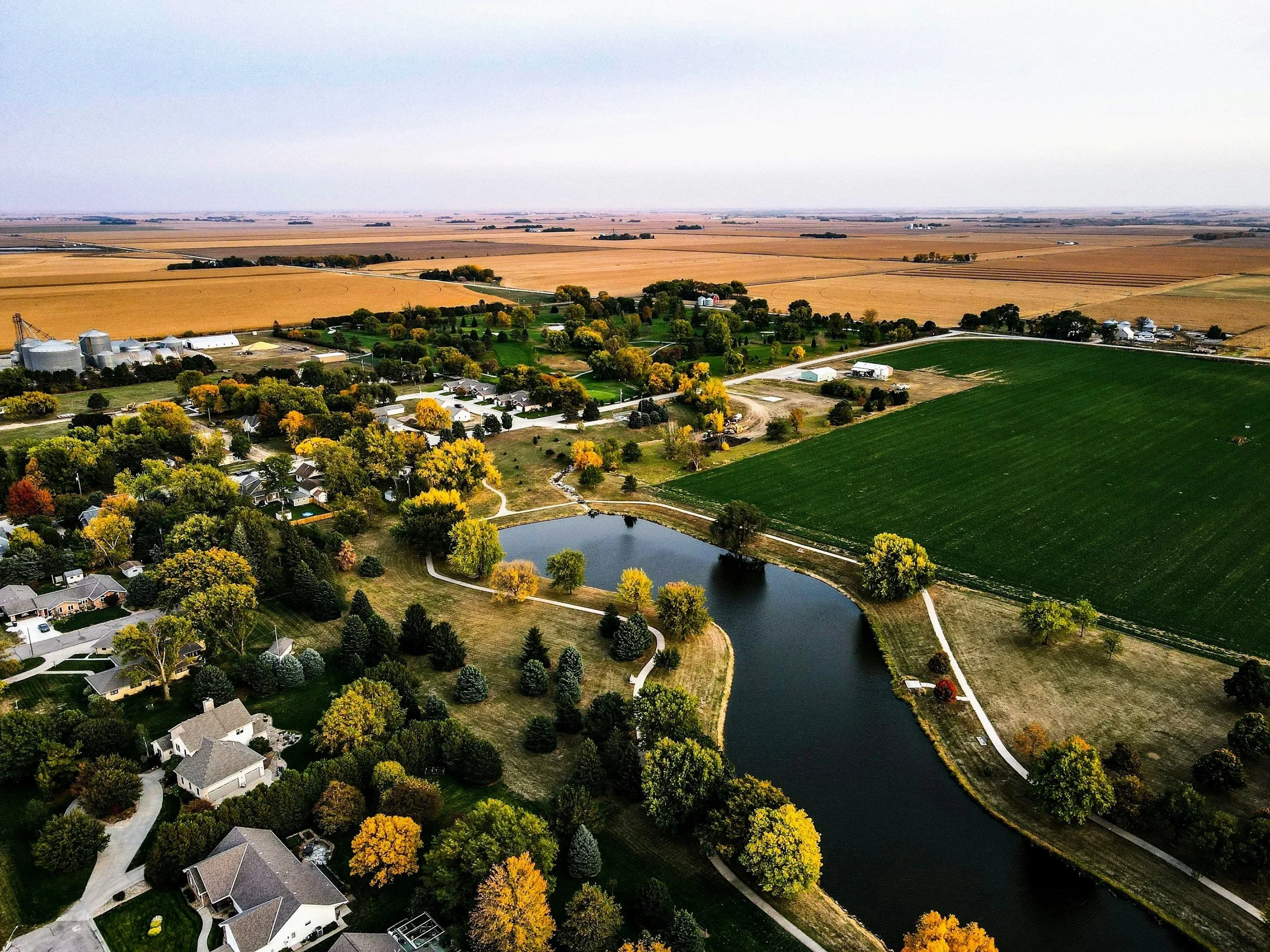 Aerial view of a countryside with a pond, houses, and farmland, showcasing fall foliage colors.