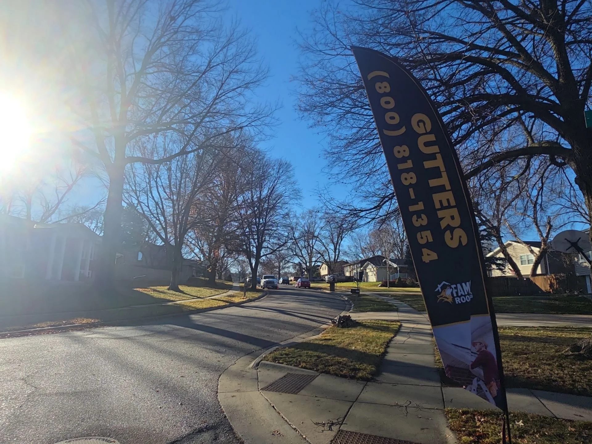 Residential neighborhood street with leafless trees, a sidewalk, and a curved road. A vertical banner with contact information is on the right side of the sidewalk, sun flare in the upper left casting light across the scene.
