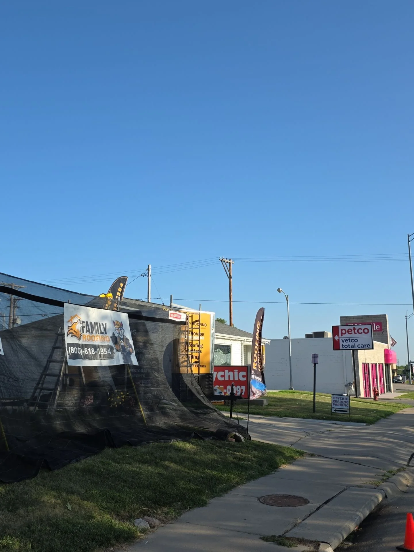 Street view showing signs for roofing companies, a Petco pet store, and a psychic reading parking sign, with a sidewalk, grass, and utility poles under a clear blue sky.