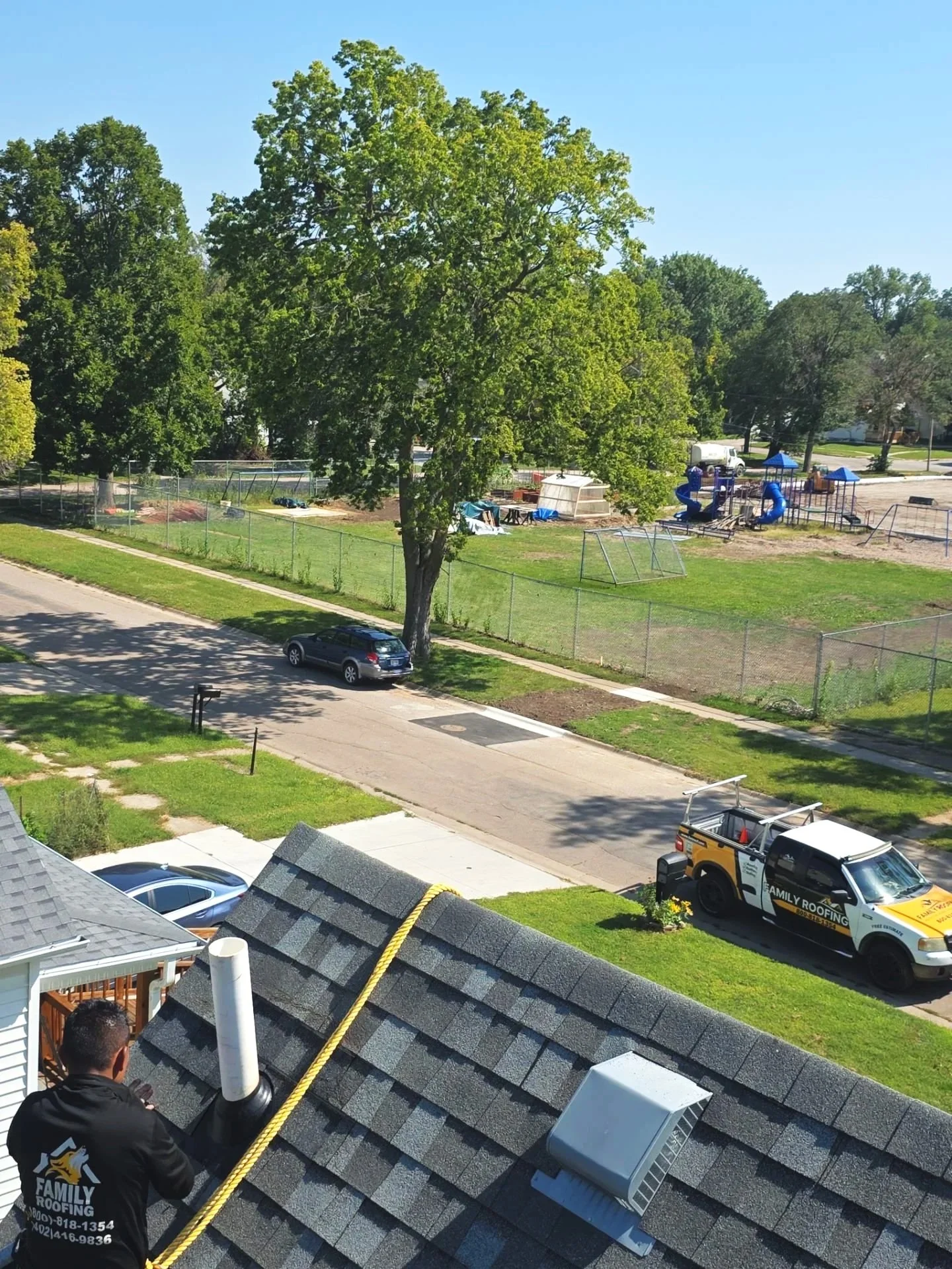 A roof with a worker installing a chimney pipe, yellow safety rope, air conditioning unit, and parked roofing company's truck, with a neighborhood street, cars, and a park with playground in the background.