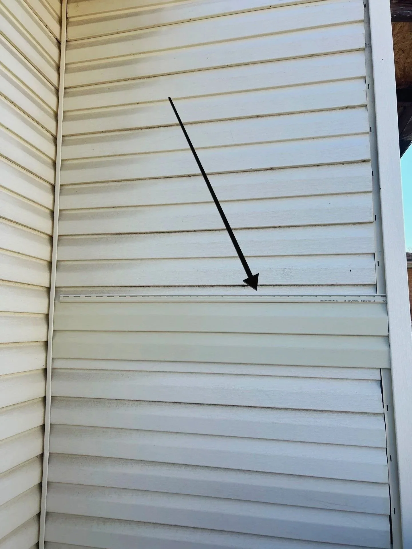 Exterior view of a beige house with vinyl siding, featuring a long metal level tool placed horizontally and marked with measurements, pointing downward.