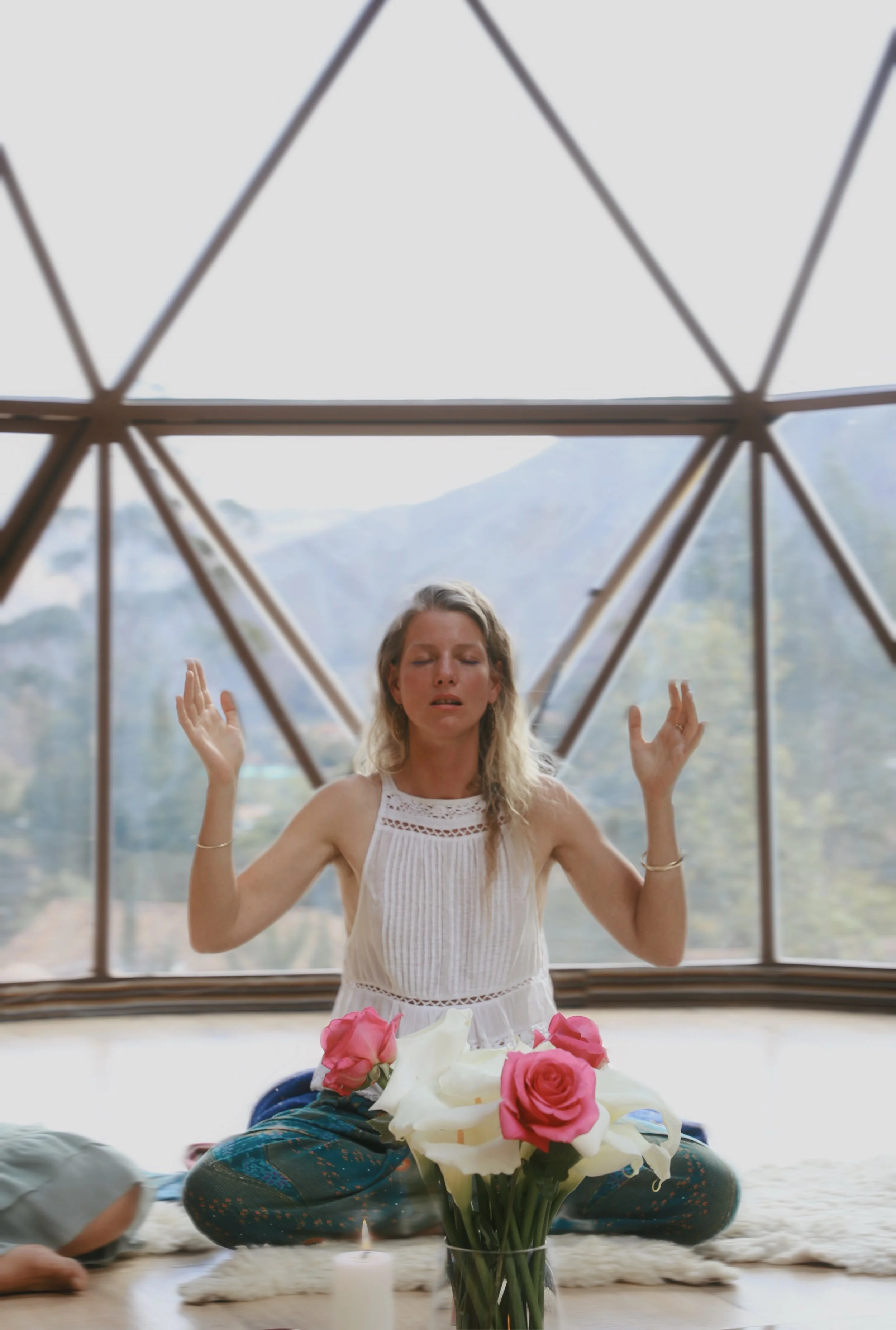 Side profile of a woman with long blonde hair, sitting in nature with mountains and greenery in the background, holding her chest with her eyes closed.