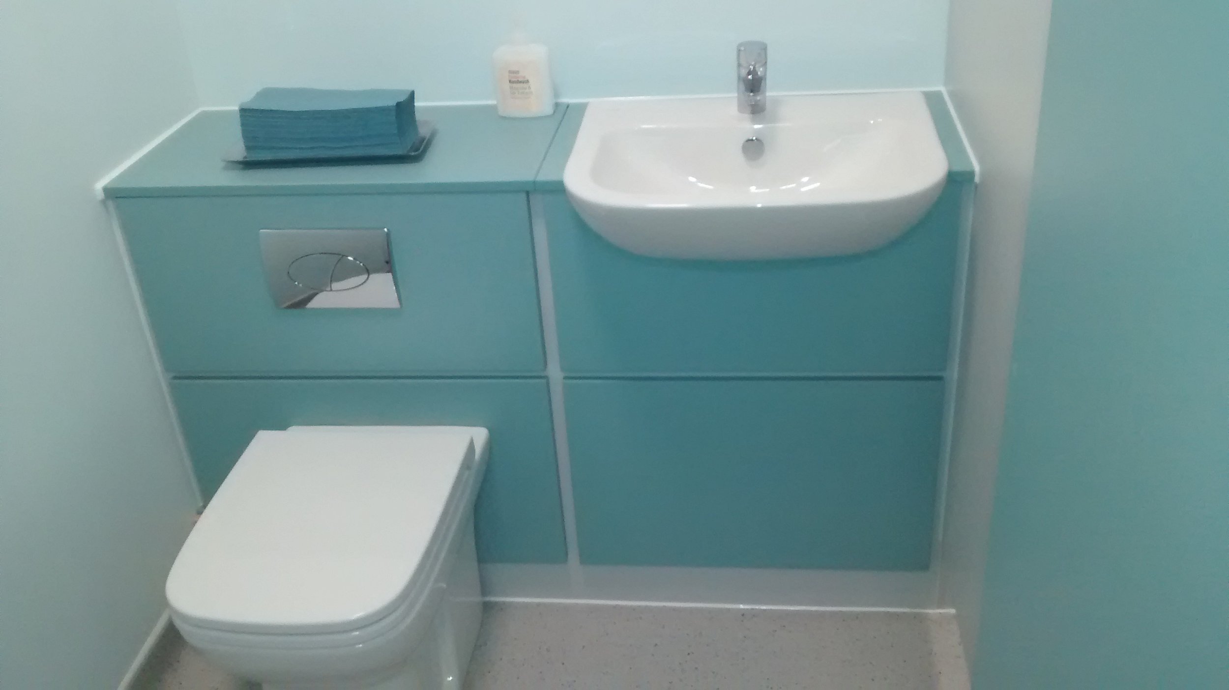 A small bathroom with a light blue cabinet, a white toilet, a small sink, and a bottle of hand sanitizer or soap on the cabinet.