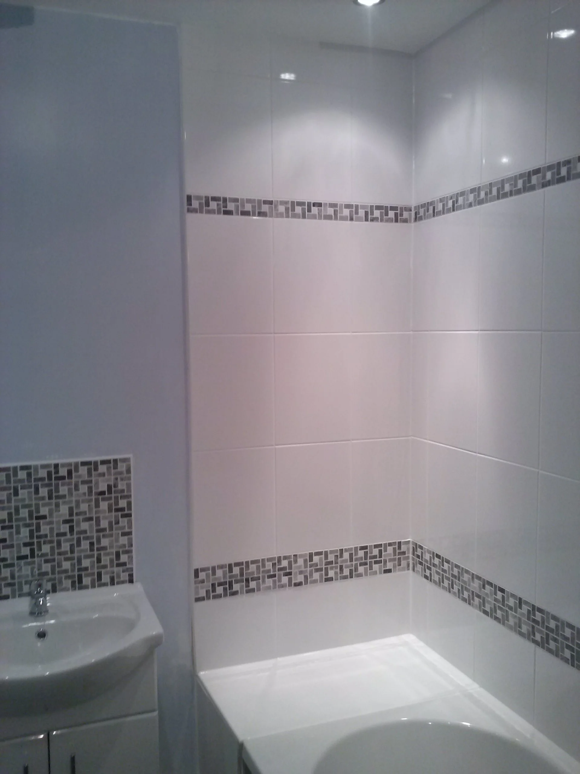 A corner of a modern bathroom with white tiled walls, gray mosaic border tiles, a small white sink, and a bathtub with a white edge.