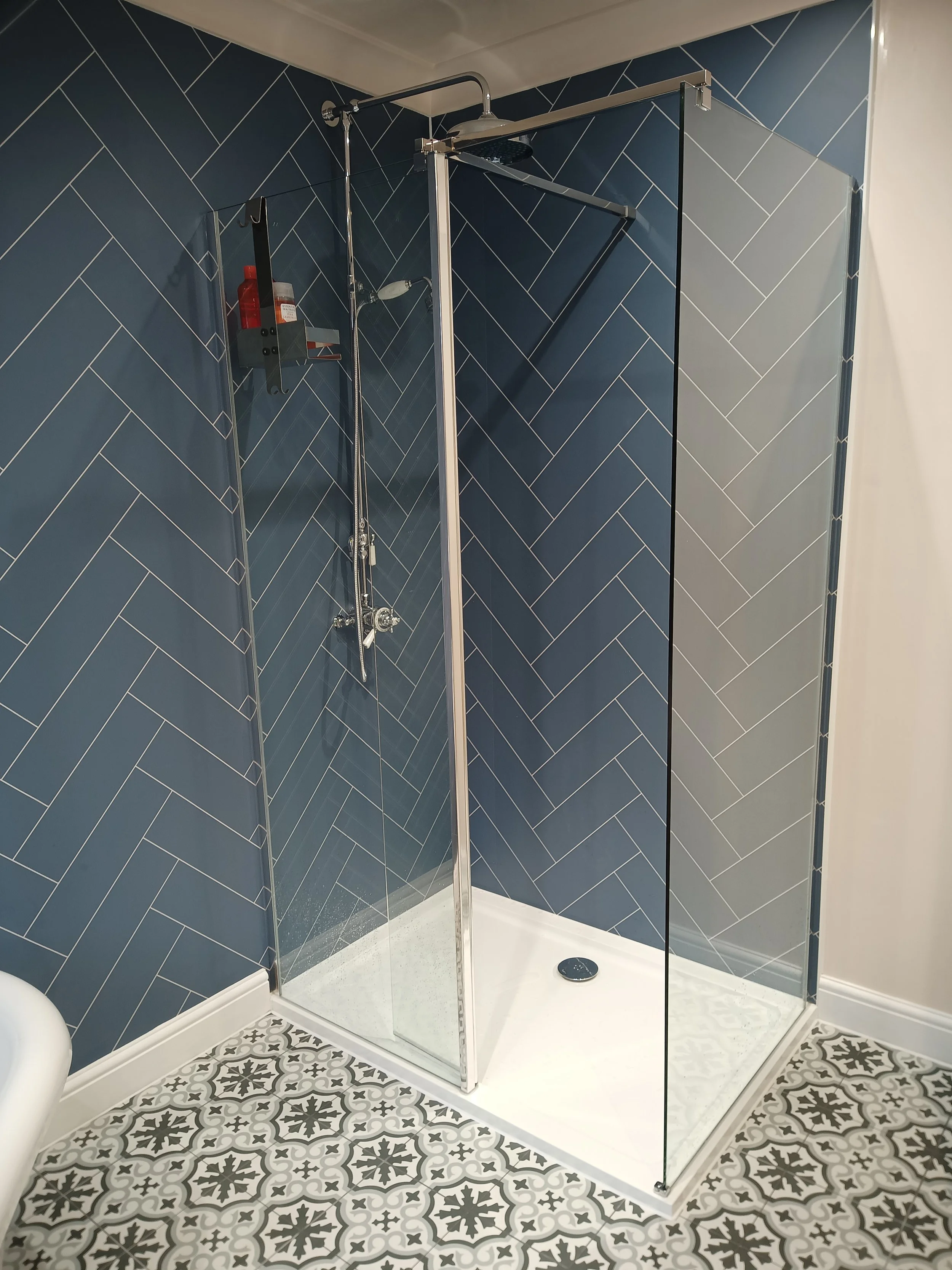 Modern shower with dark blue tile walls in a herringbone pattern, a clear glass enclosure, and patterned floor tiles.