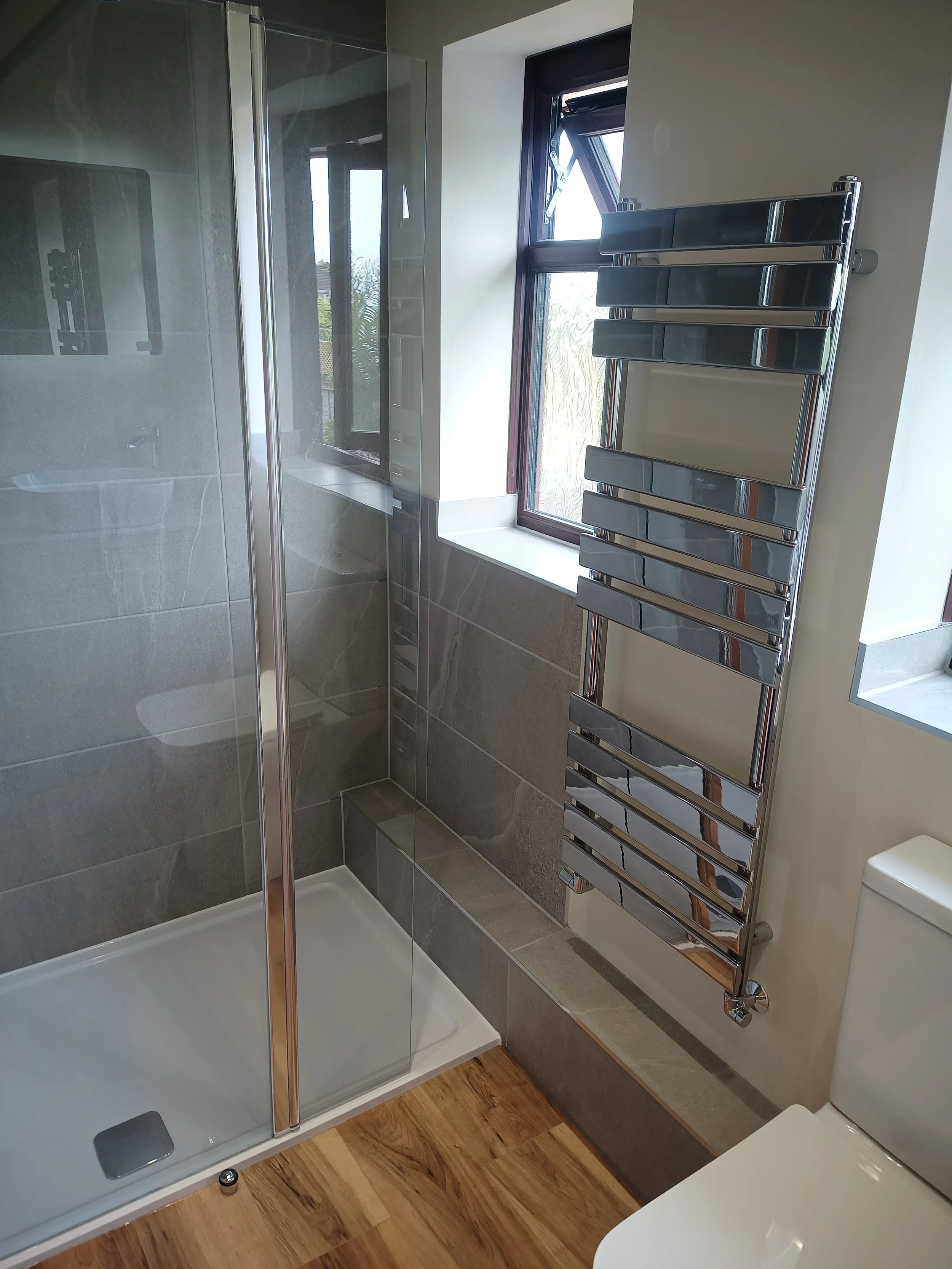 Modern bathroom features a glass shower enclosure, a chrome towel radiator, a window with a black frame, and a white toilet, with wooden flooring and grey wall tiles.