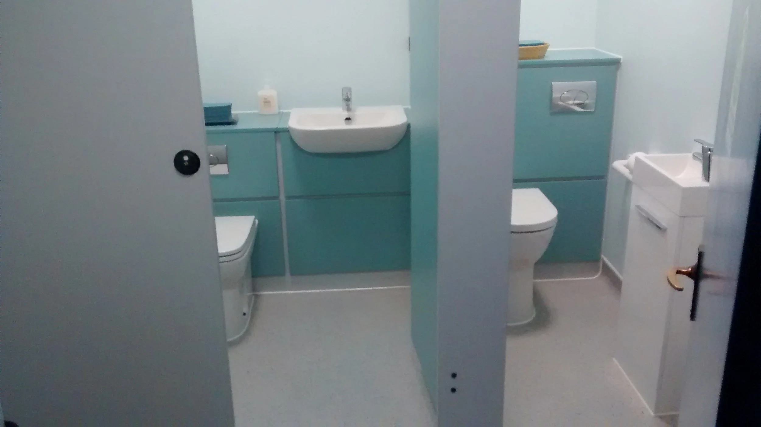 A small hospital or clinic bathroom with a blue and white color scheme, containing a toilet, a small sink, and a high-mounted toilet flush panel.