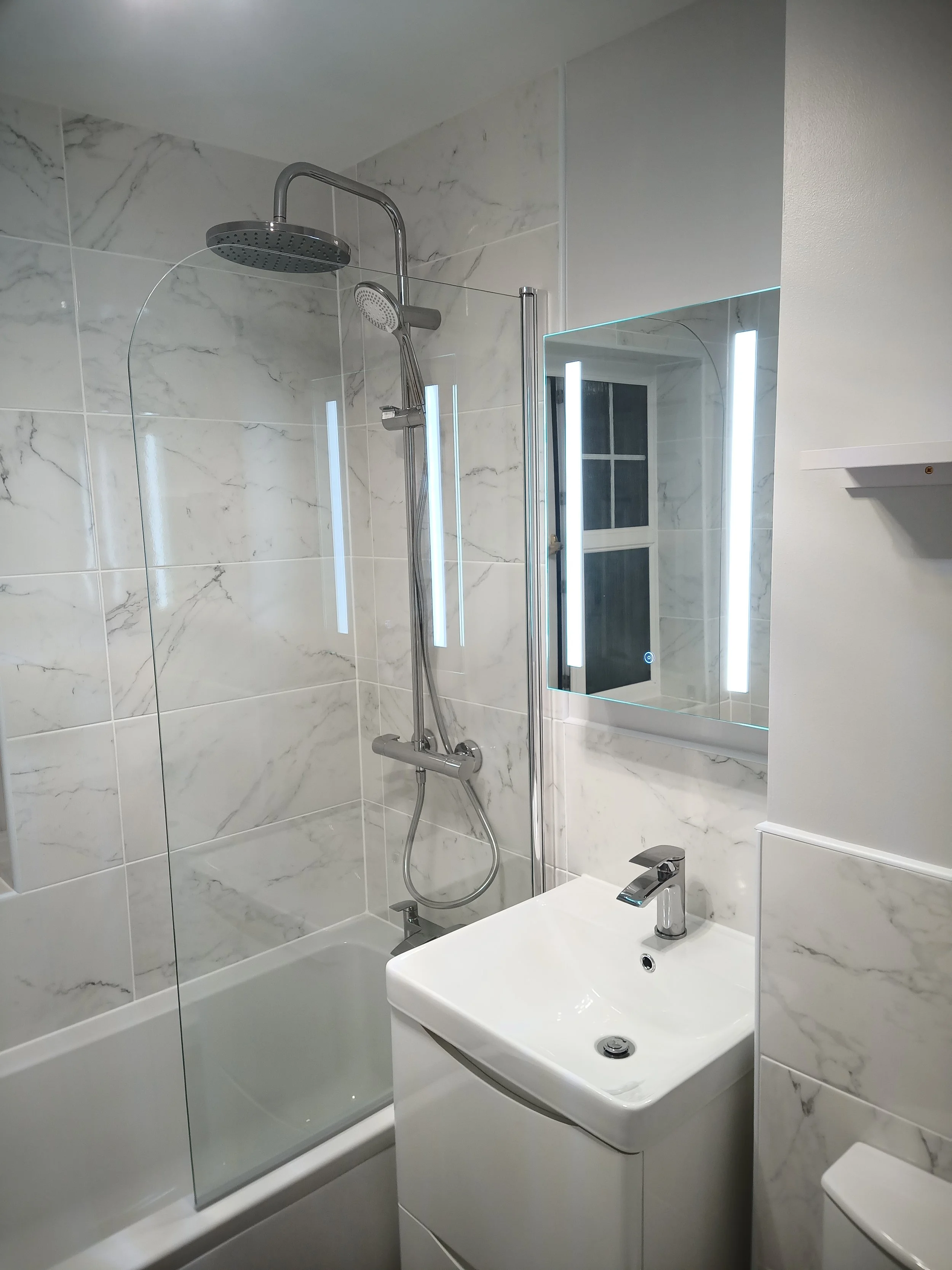 Modern bathroom with a shower area that has a glass panel, a small white sink with a chrome faucet, and a mirror with vertical LED lights, marble tile walls, and a window.