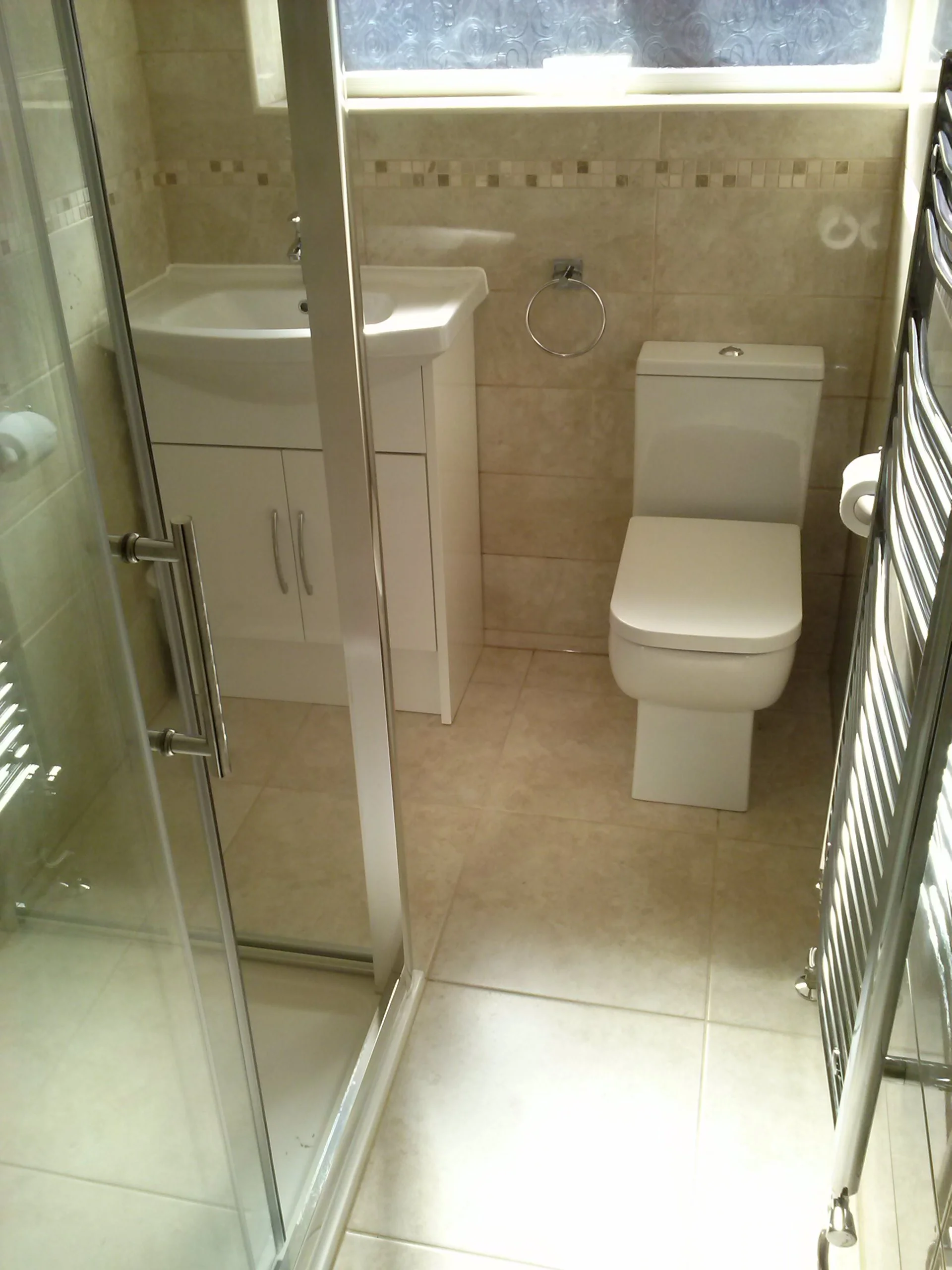 Small bathroom with beige tiled walls and floor, featuring a glass shower enclosure, white vanity with sink, toilet, towel ring, and heated towel rail.