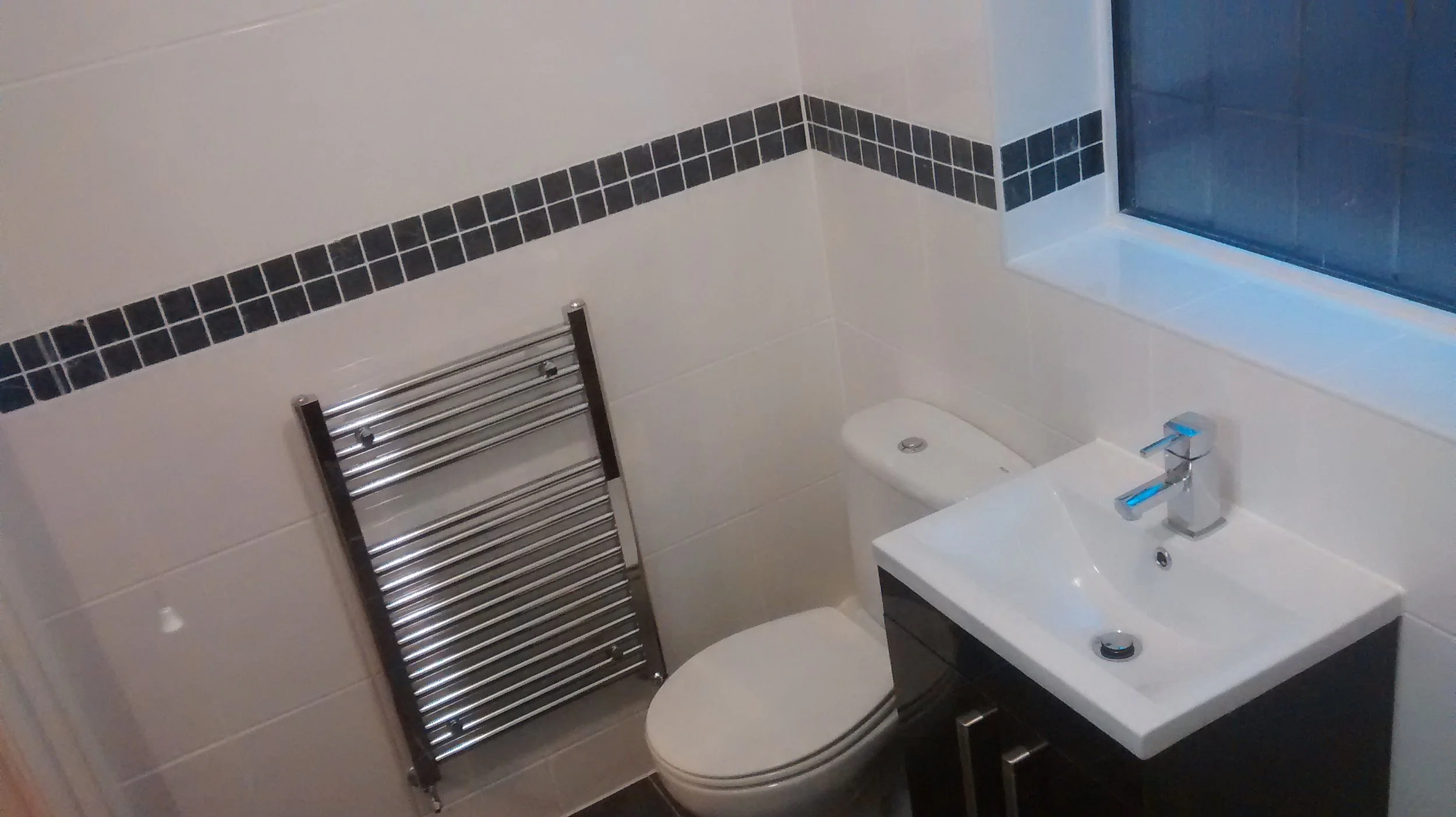 Small bathroom with a white toilet, a black vanity with a white sink and silver faucet, a chrome heated towel rack, and a window above the sink.
