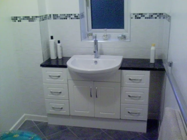 Bathroom sink with white cabinet, silvery handles, black countertop, three toilet paper rolls on the left, a yellow bottle on the right, and a small window above.