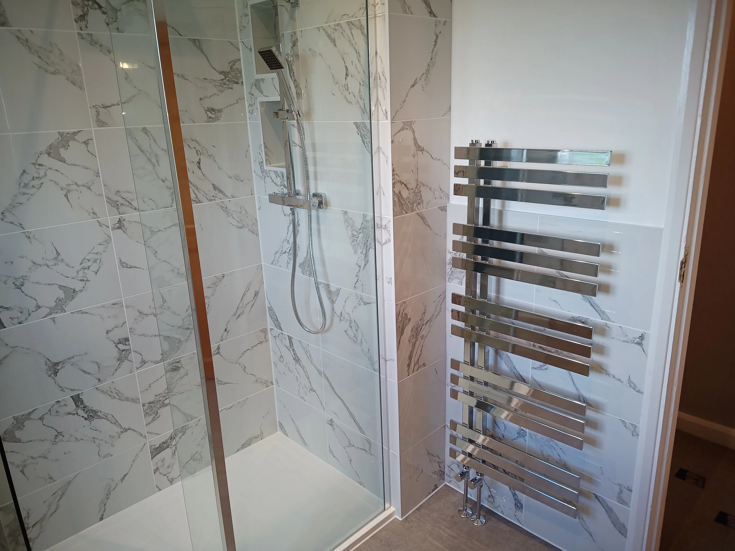 A modern bathroom shower with marble tile walls and a glass door, alongside a chrome towel radiator.