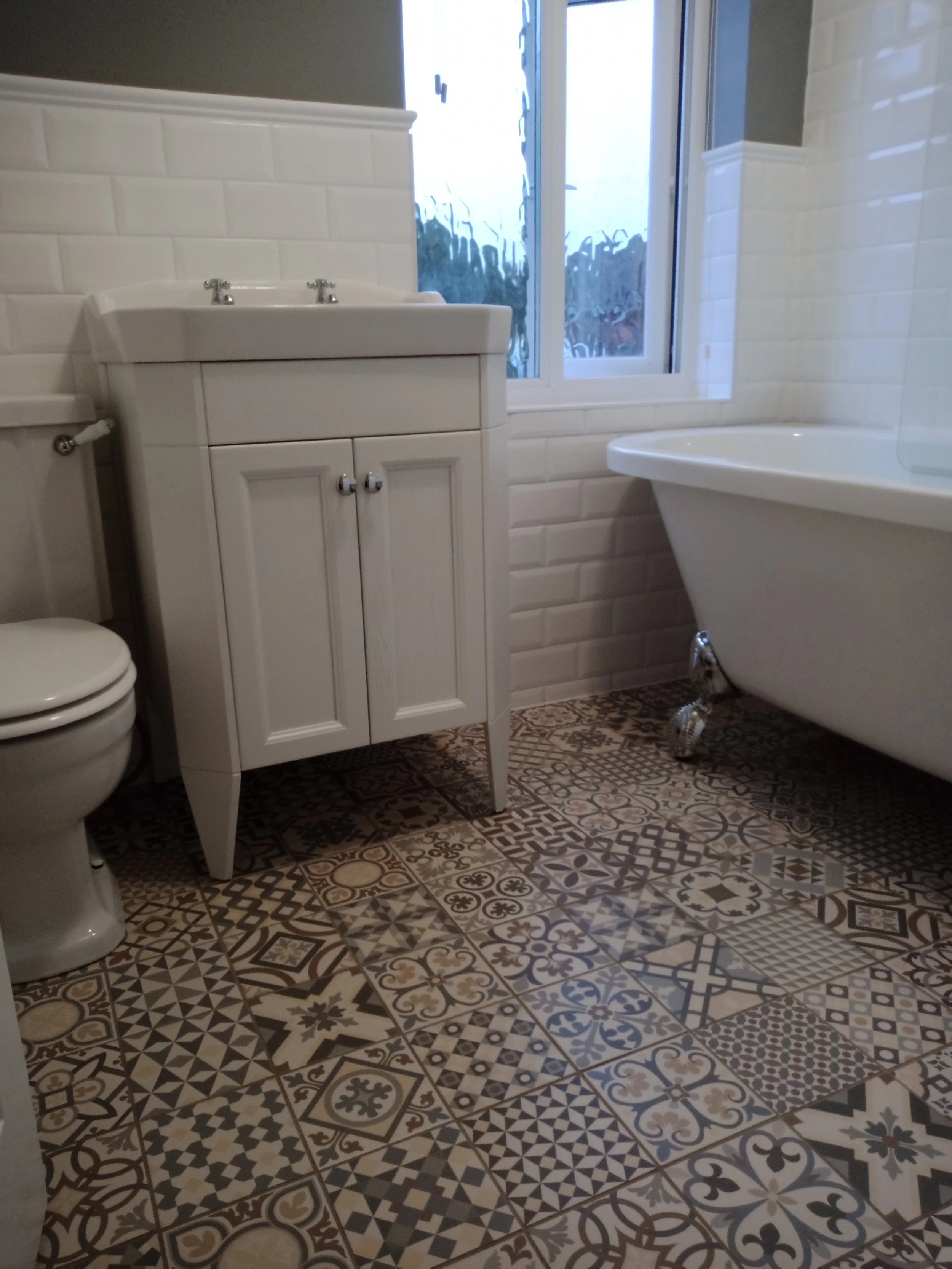 A bathroom with patterned tile floor, white bathtub, white sink cabinet, toilet, and window with water droplets on the glass.