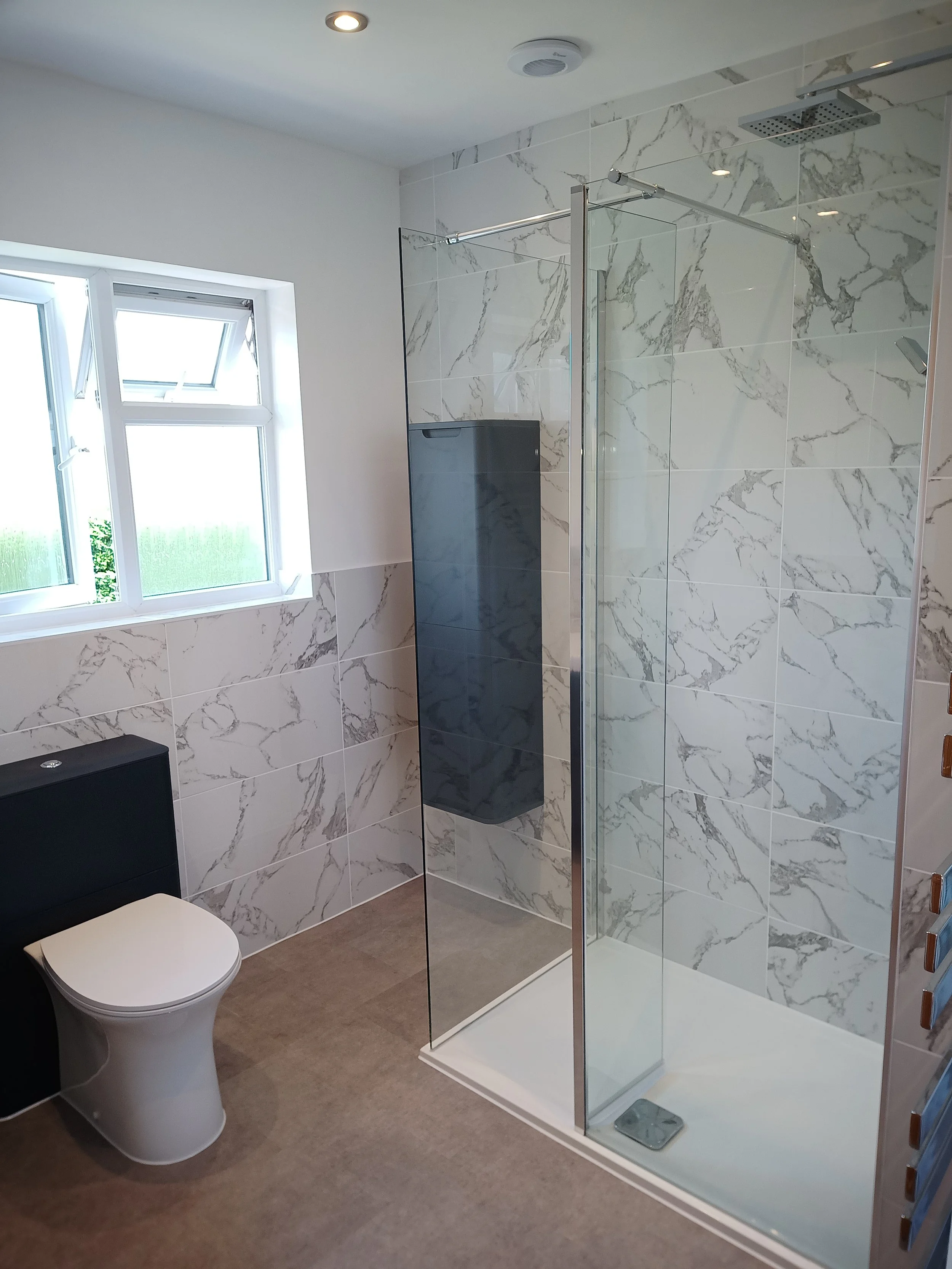 Modern bathroom featuring a white toilet, a black storage cabinet, a window, and a walk-in shower with marble-patterned tiles and a glass door.