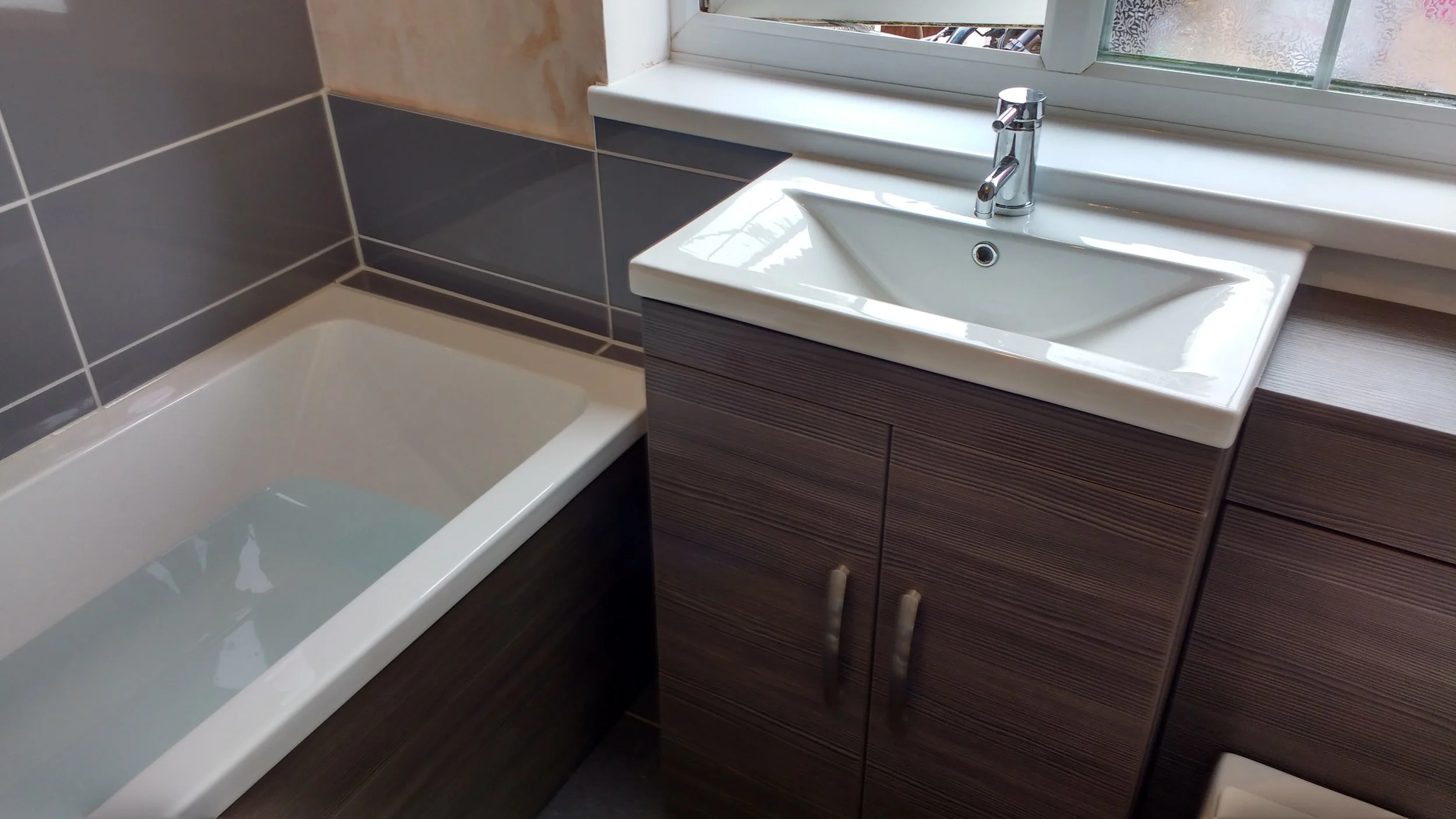 Bathroom with bathtub filled with water, dark wooden vanity with a white sink and a chrome faucet, and a window above the sink.