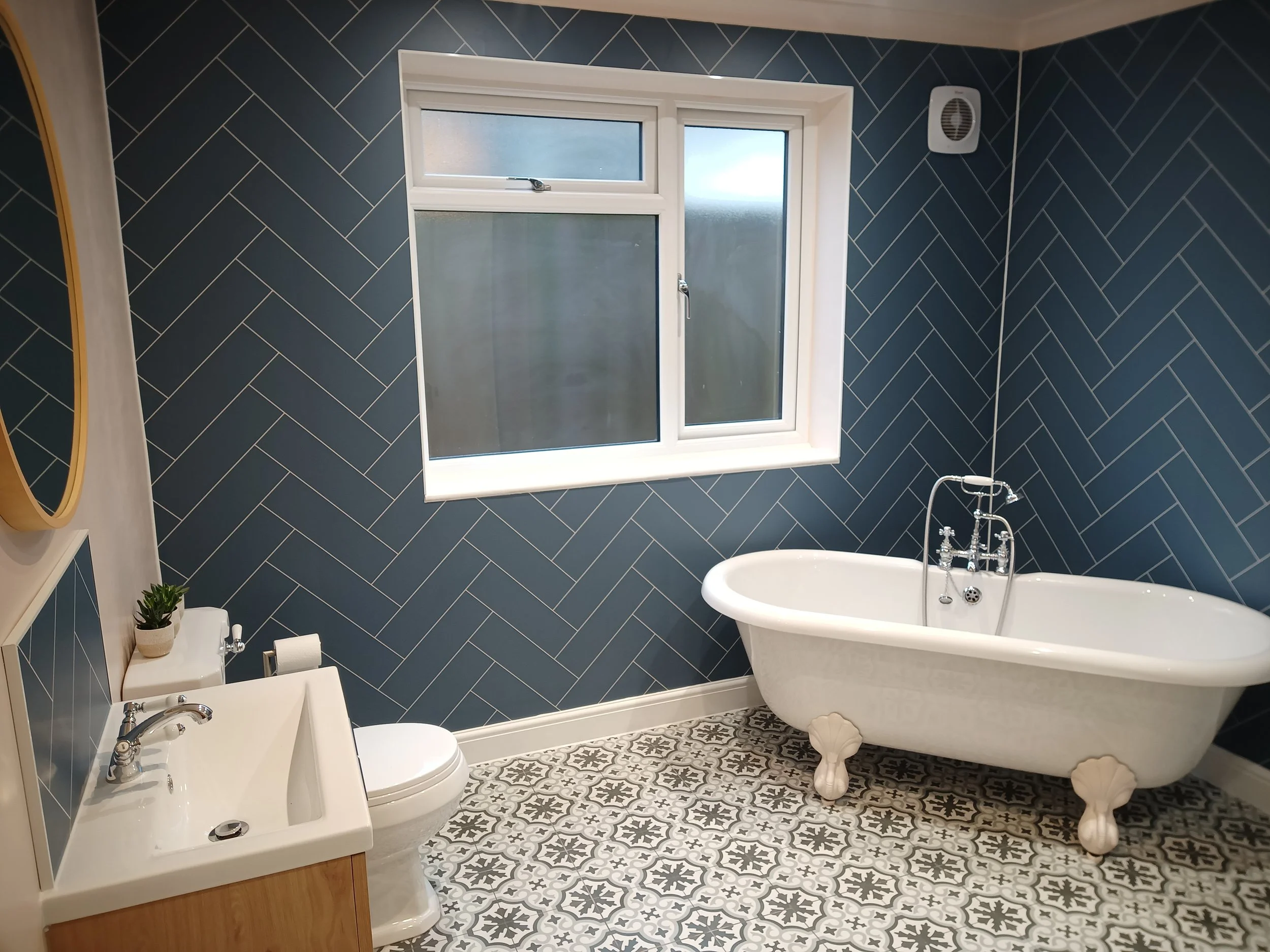 Bathroom with blue tiled walls, a white clawfoot bathtub, a window, a small sink, and a toilet. Decorative patterned floor tiles.