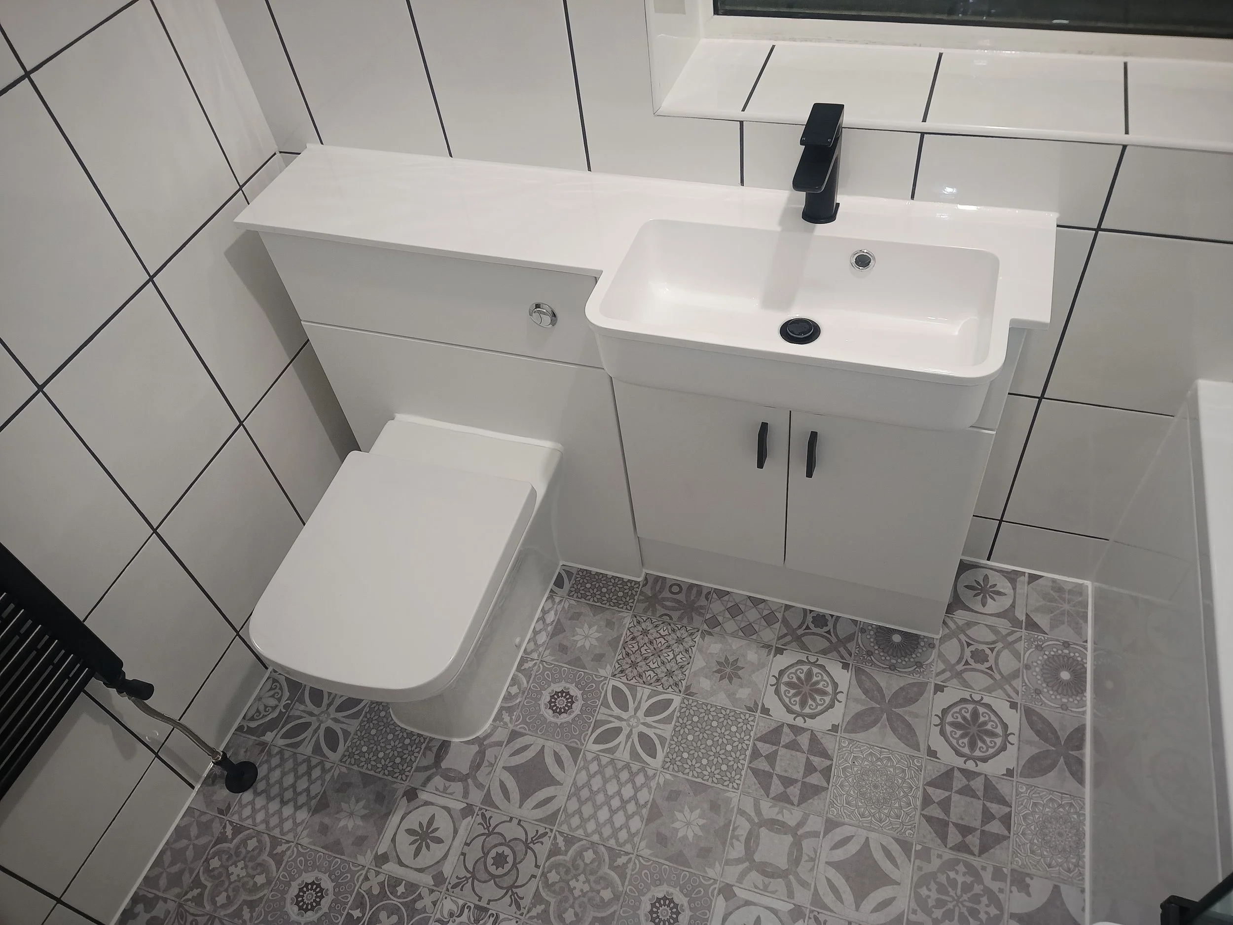 Modern bathroom with white tiles and geometric-patterned floor tiles, featuring a wall-mounted toilet, a small white vanity with a sink, and a mirror.