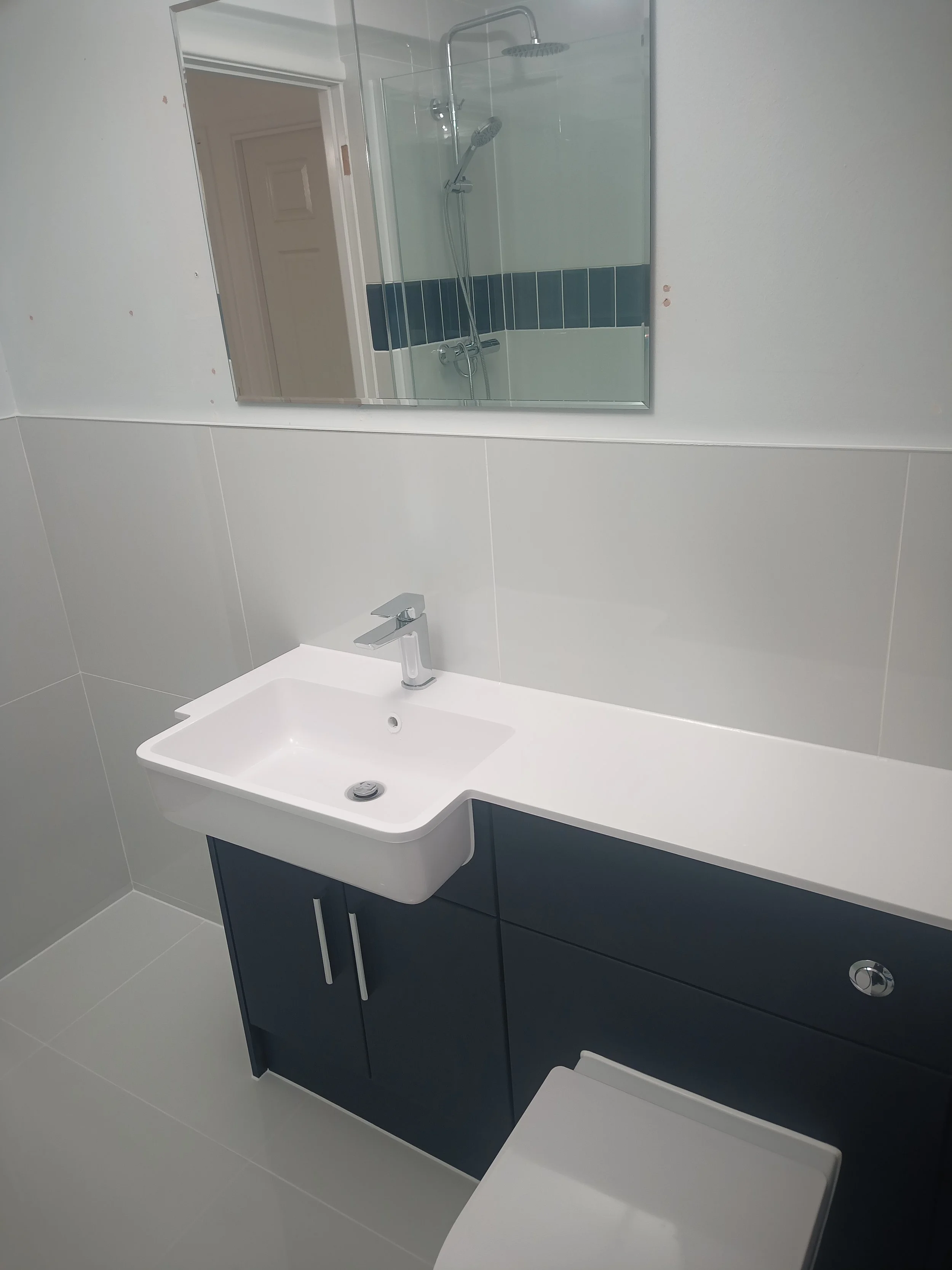 Modern bathroom with white sink, navy blue cabinet, large mirror, and shower with glass door in the background.
