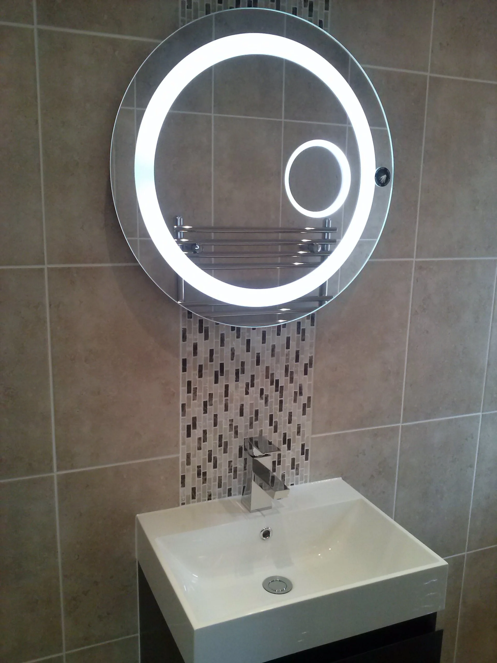 Modern bathroom mirror with built-in LED light above a white rectangular sink with a chrome faucet, mounted on a tiled wall with beige and mosaic tiles.