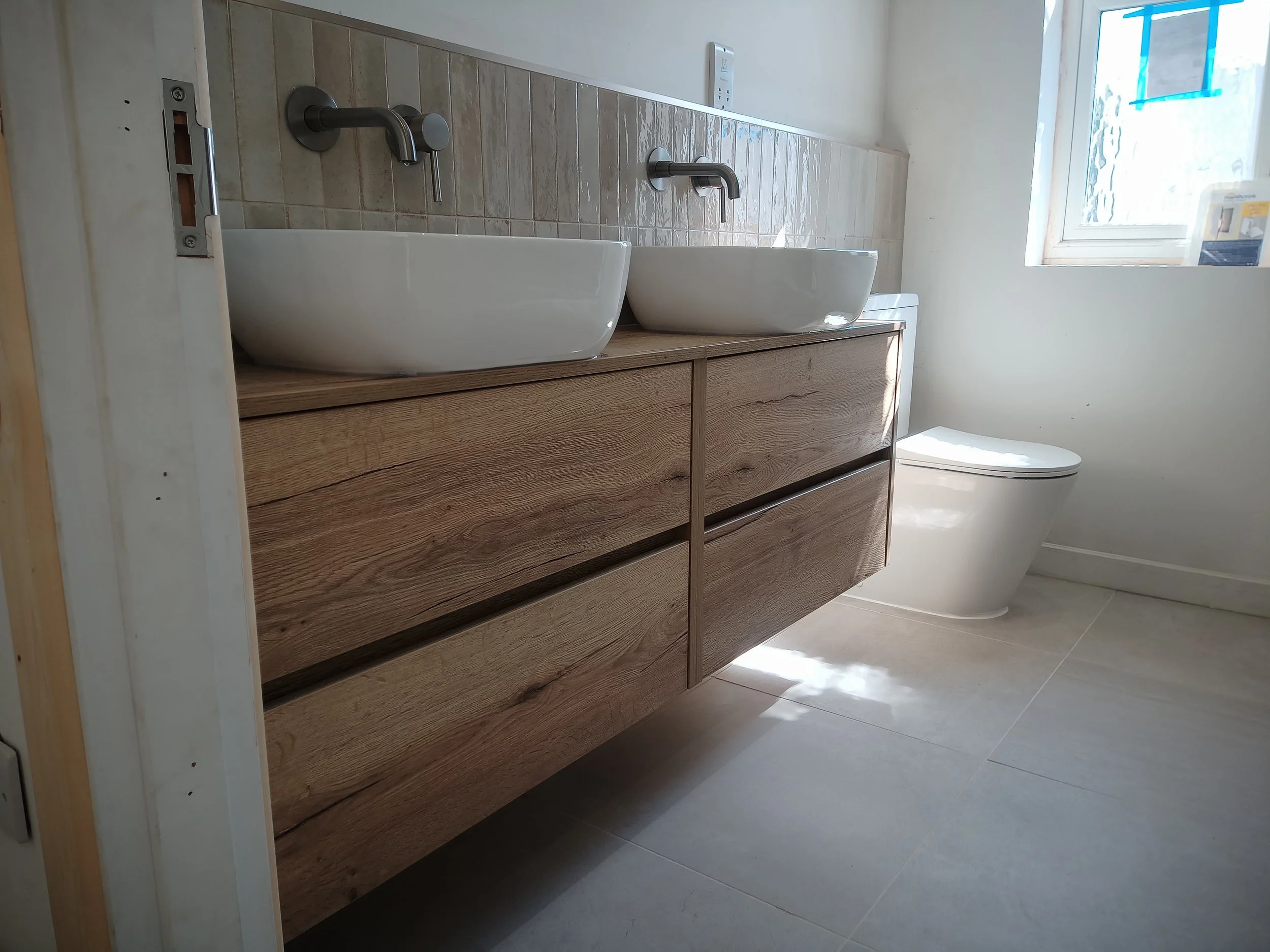 Bathroom with two vessel sinks on a wooden vanity, modern wall-mounted faucets, a toilet, and a window letting in natural light.