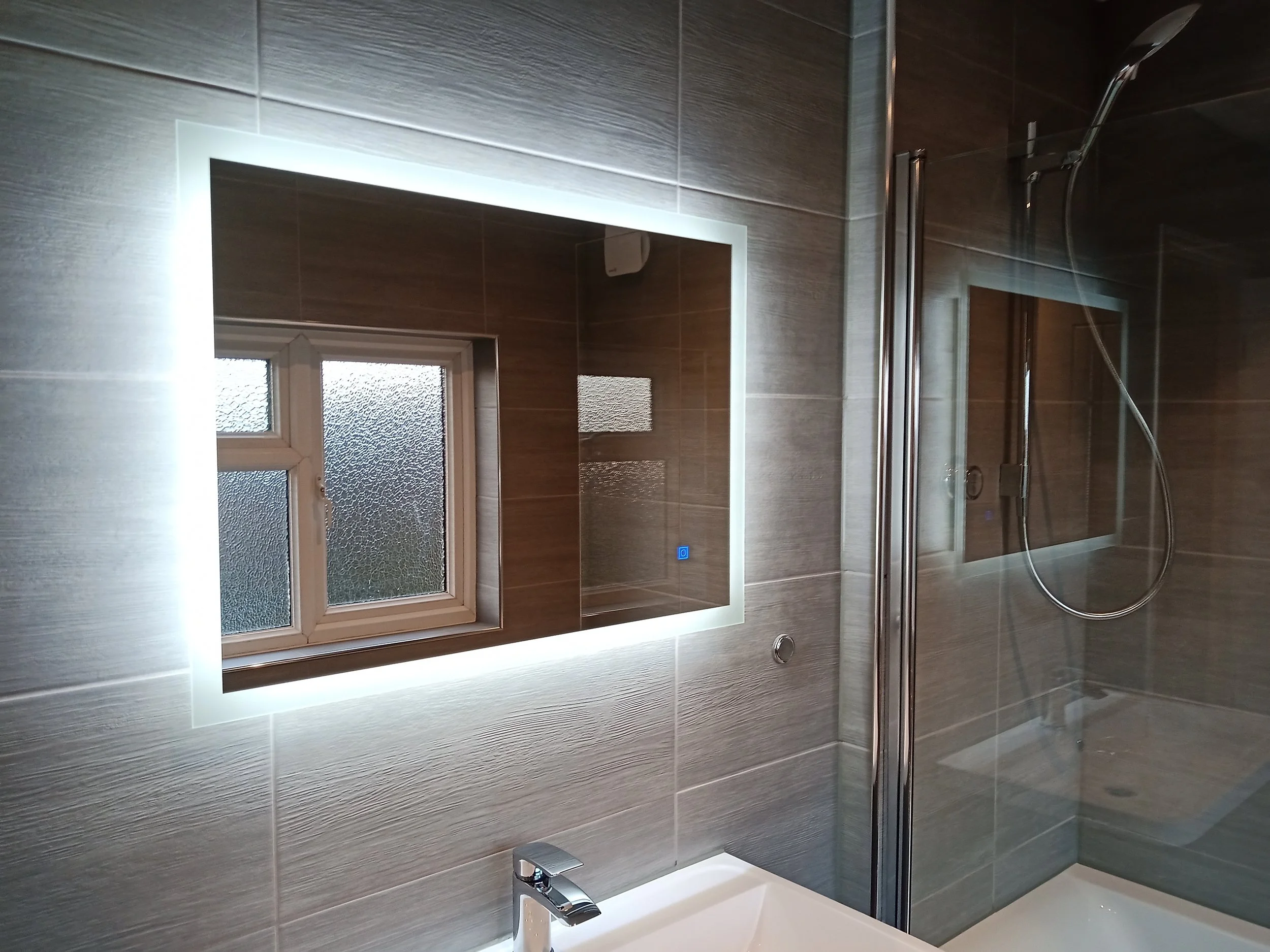 Modern bathroom with a backlit mirror, a textured wall, a small frosted window, and a glass shower enclosure with a showerhead.
