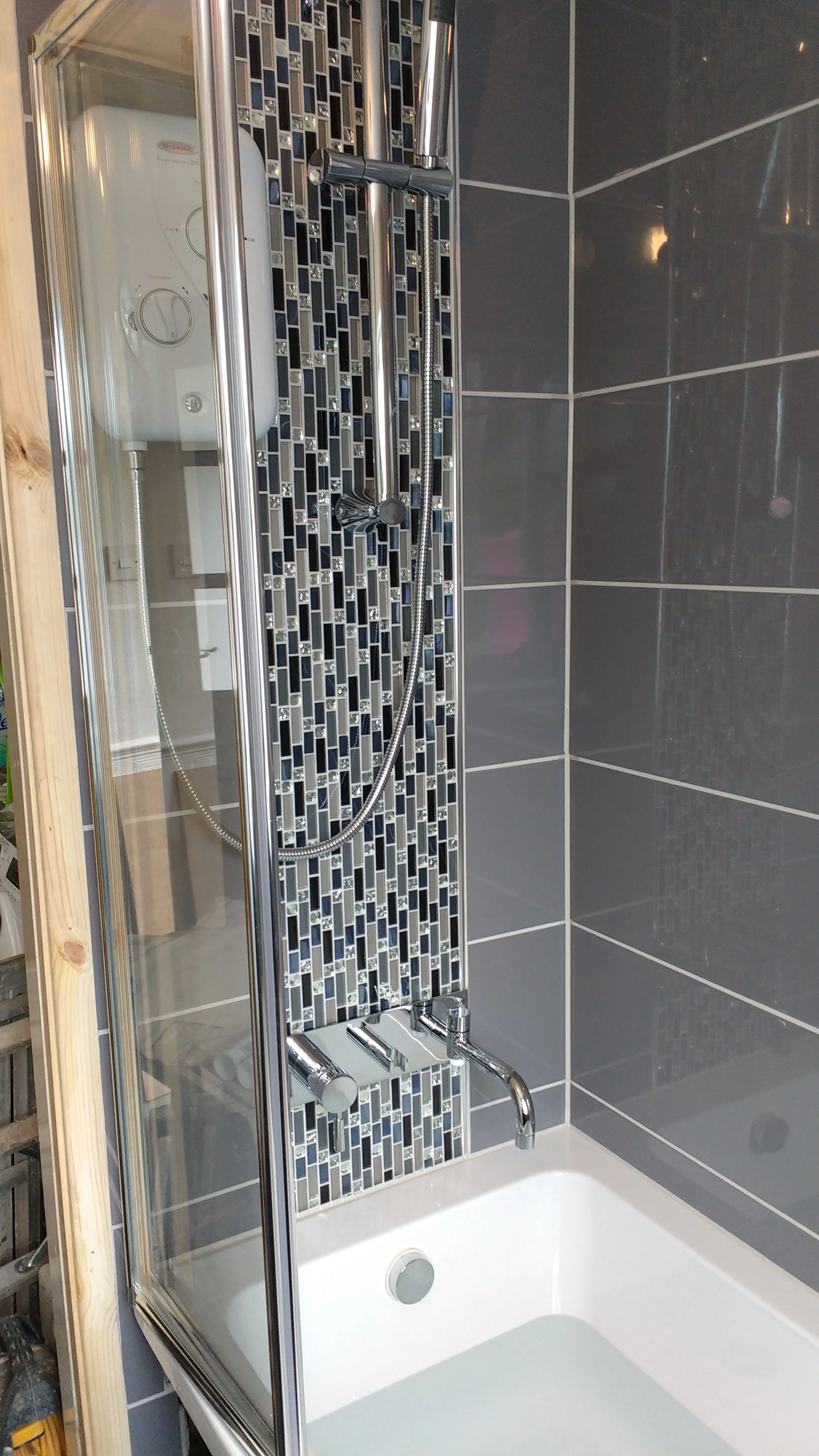 Bathroom shower with dark gray tile walls, a small mosaic tile strip, and a glass door with chrome handle, featuring a built-in bathtub and a handheld showerhead.