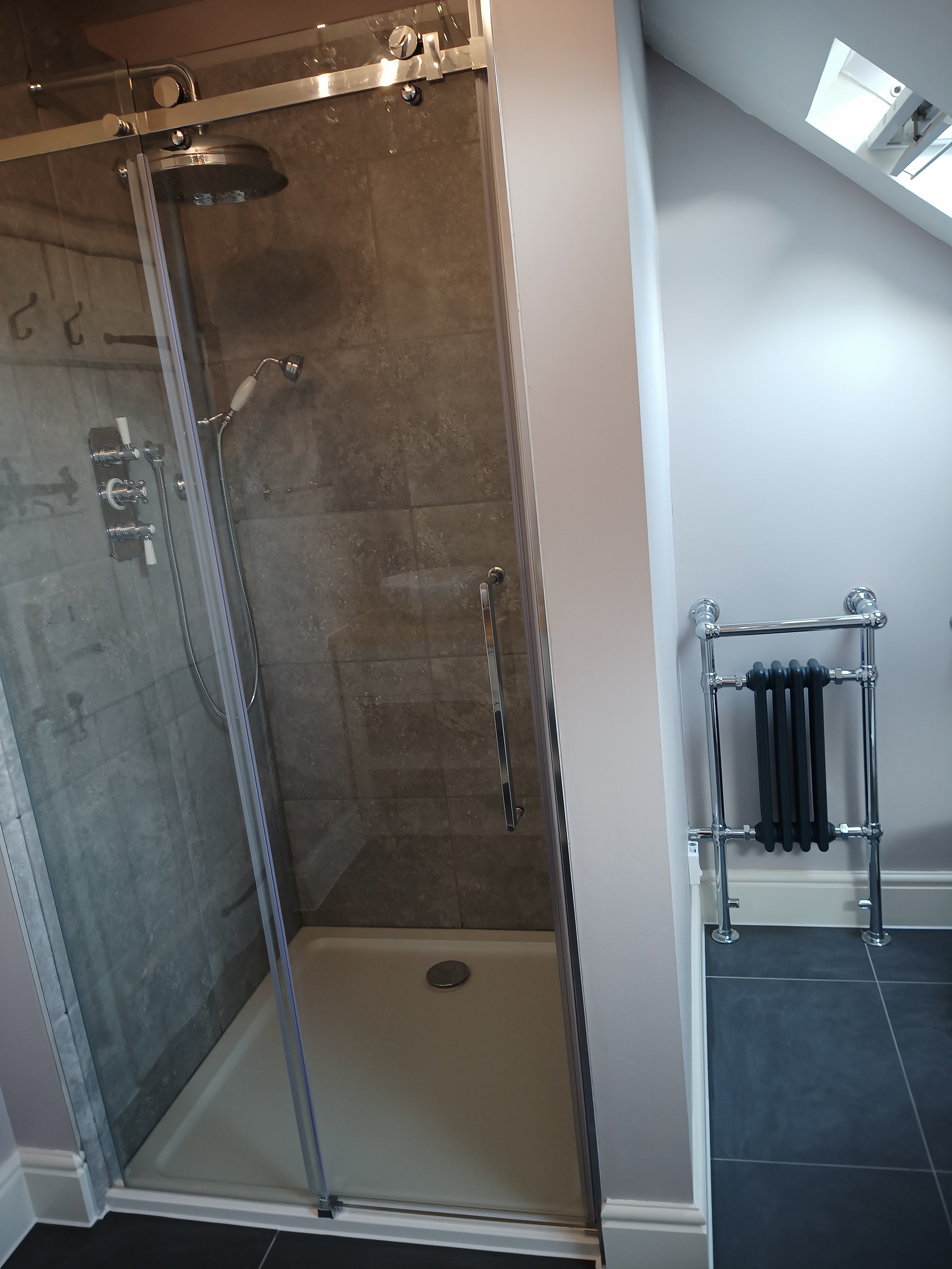 A modern bathroom features a glass-enclosed shower with large brown tiles, a chrome showerhead and controls, and a white shower tray. Next to the shower is a wall-mounted radiator with black towel hooks in a space with dark tiled flooring and white w