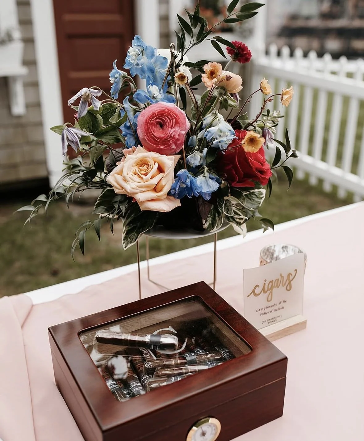 A floral arrangement with pink, cream, red, and blue flowers on a table with a wooden cigar box and a sign for cigars.