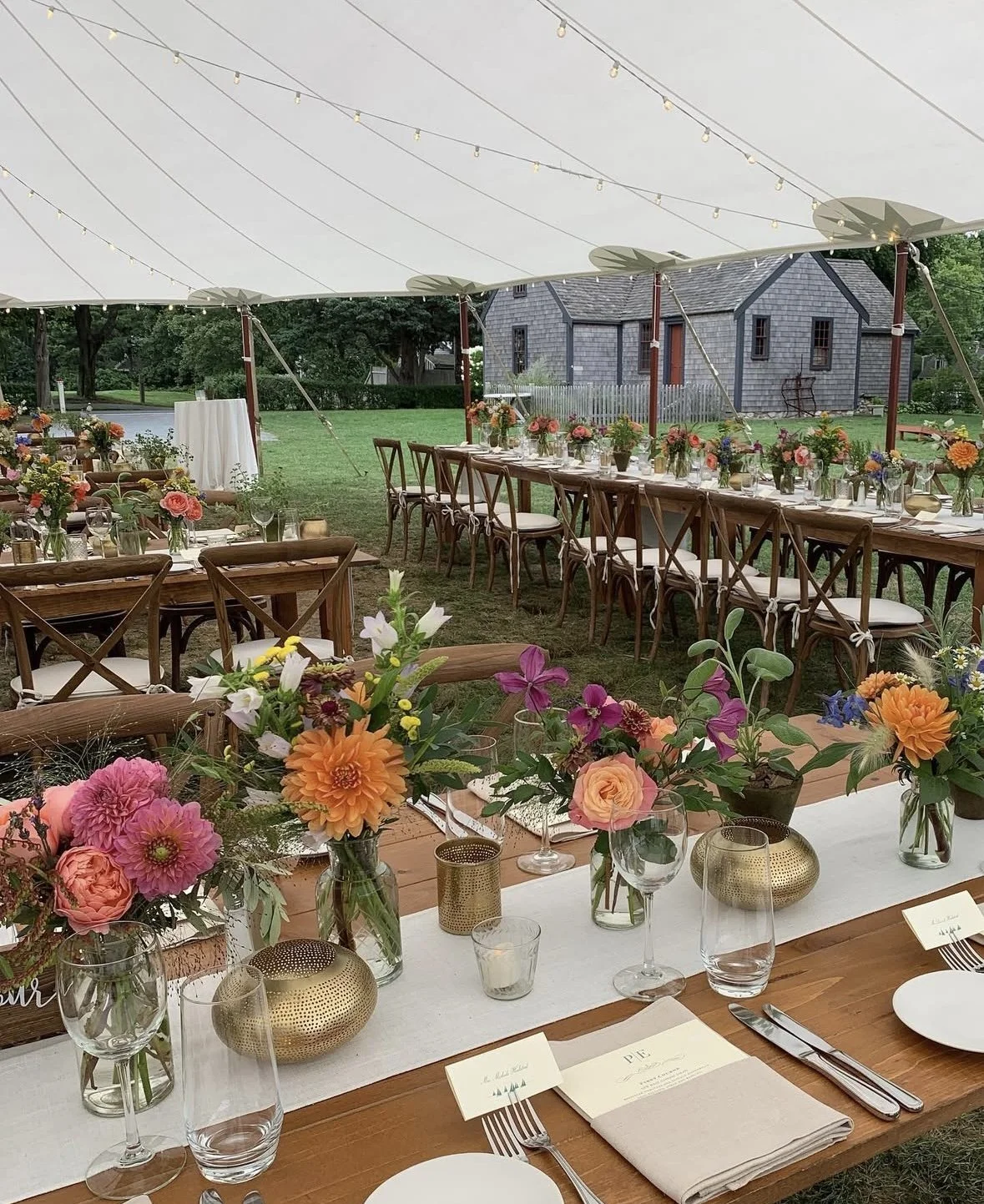 Decorated outdoor dining area under a large white canopy with long tables, floral arrangements, and string lights, set on a grassy lawn with trees and a gray wooden building in the background.