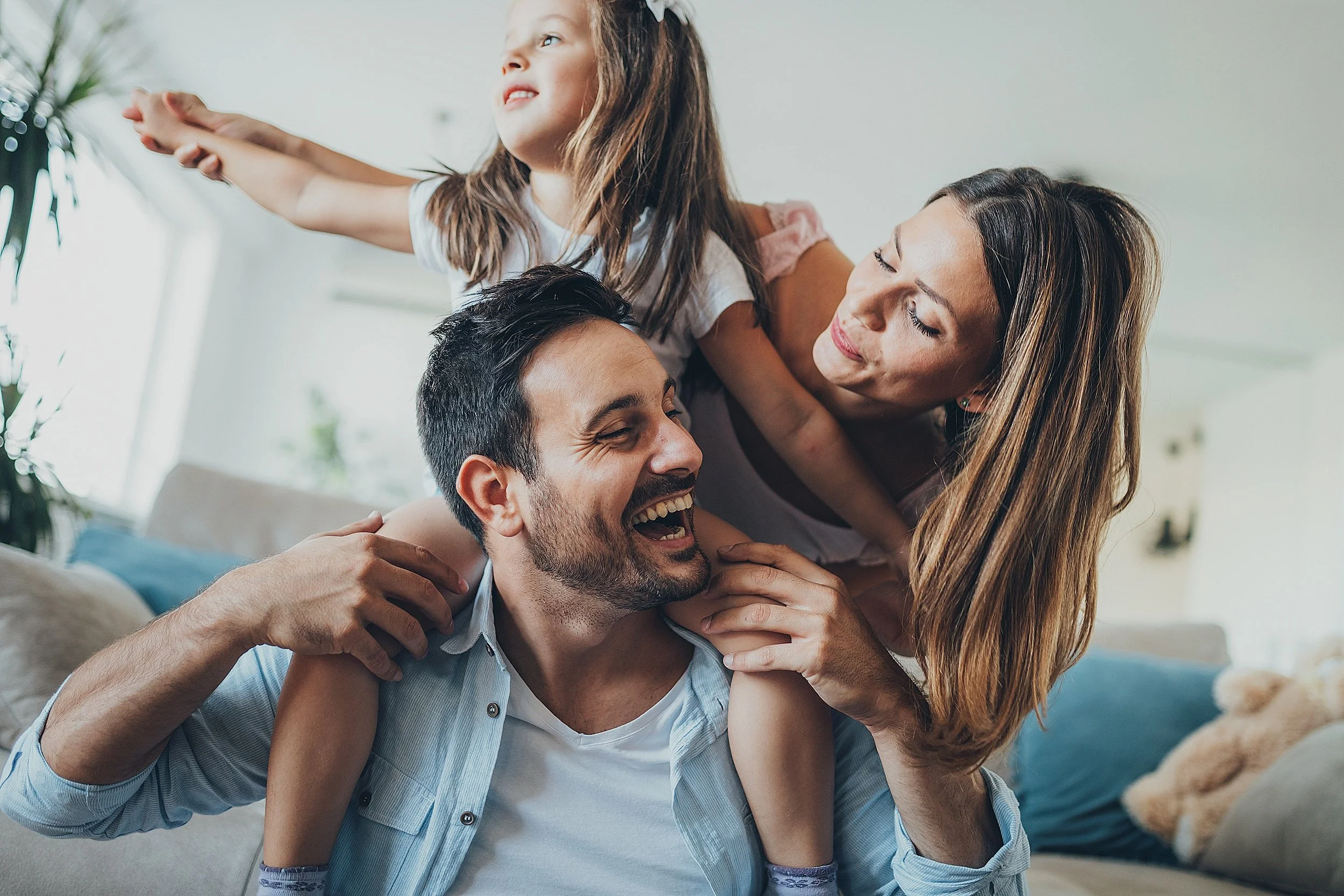 A family of three, a father, mother, and young daughter, enjoying playtime together on the sofa in their living room. The father is smiling while carrying the daughter on his shoulders, and the mother is leaning in, holding her daughter.