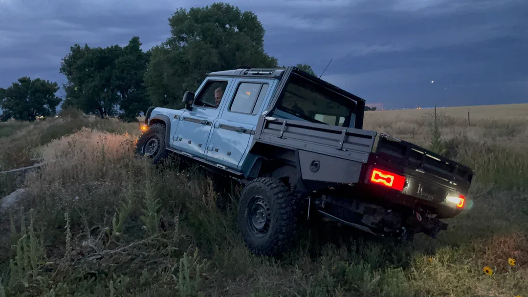 A blue pickup truck is stuck on a grassy hillside during dusk with cloudy skies.