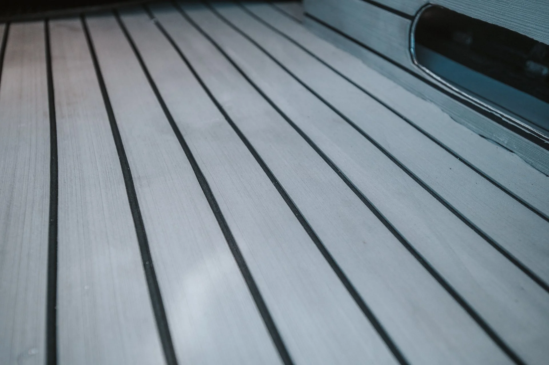 Close-up view of a boat deck with parallel wooden or composite planks with dark lines, featuring a ventilation opening on the right side.