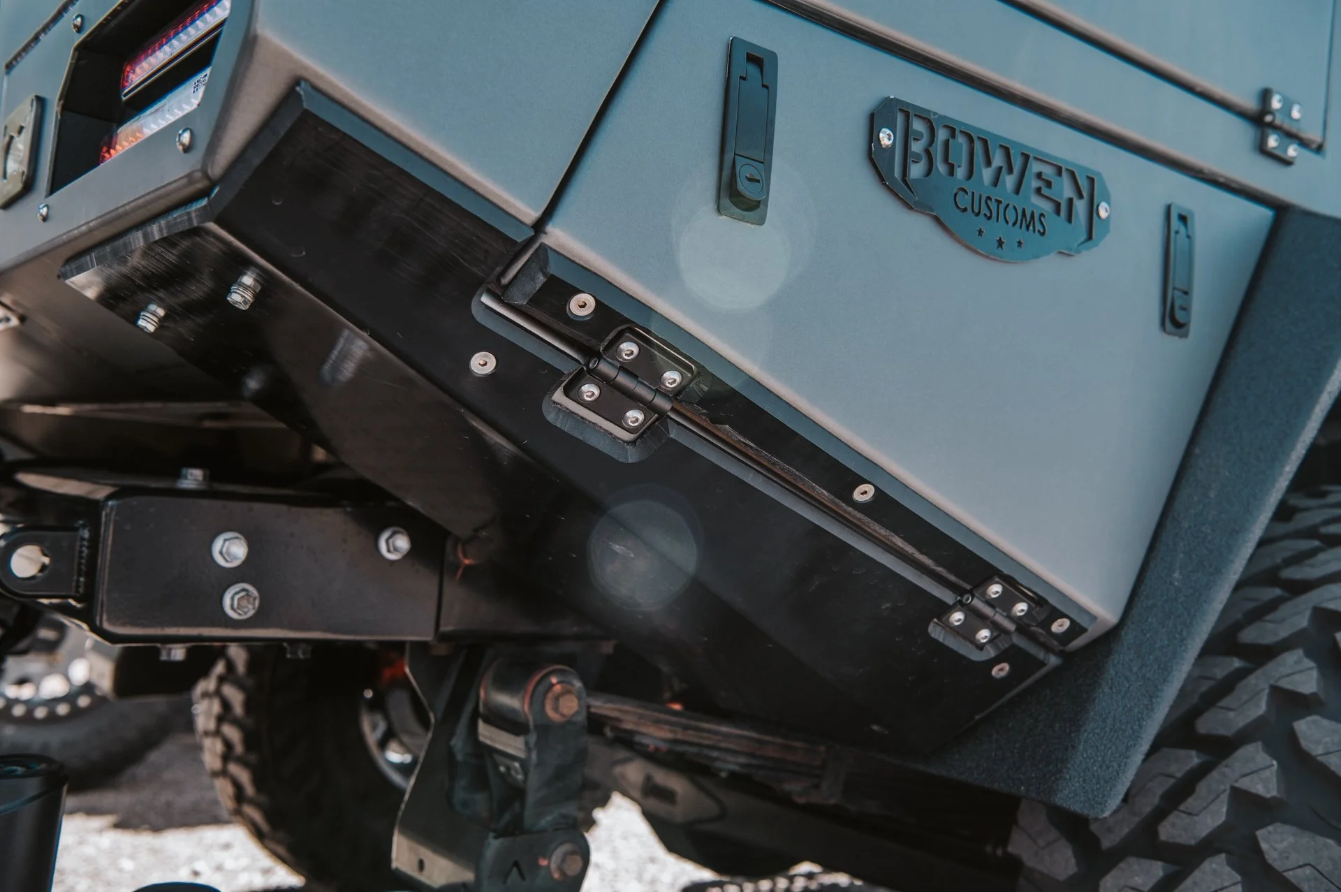 Close-up of a blue vehicle trunk or cargo area with a metal badge that reads 'BOWEN CUSTOMS,' showing a handle latch and hinges, with parts of a rugged tire visible at the bottom.
