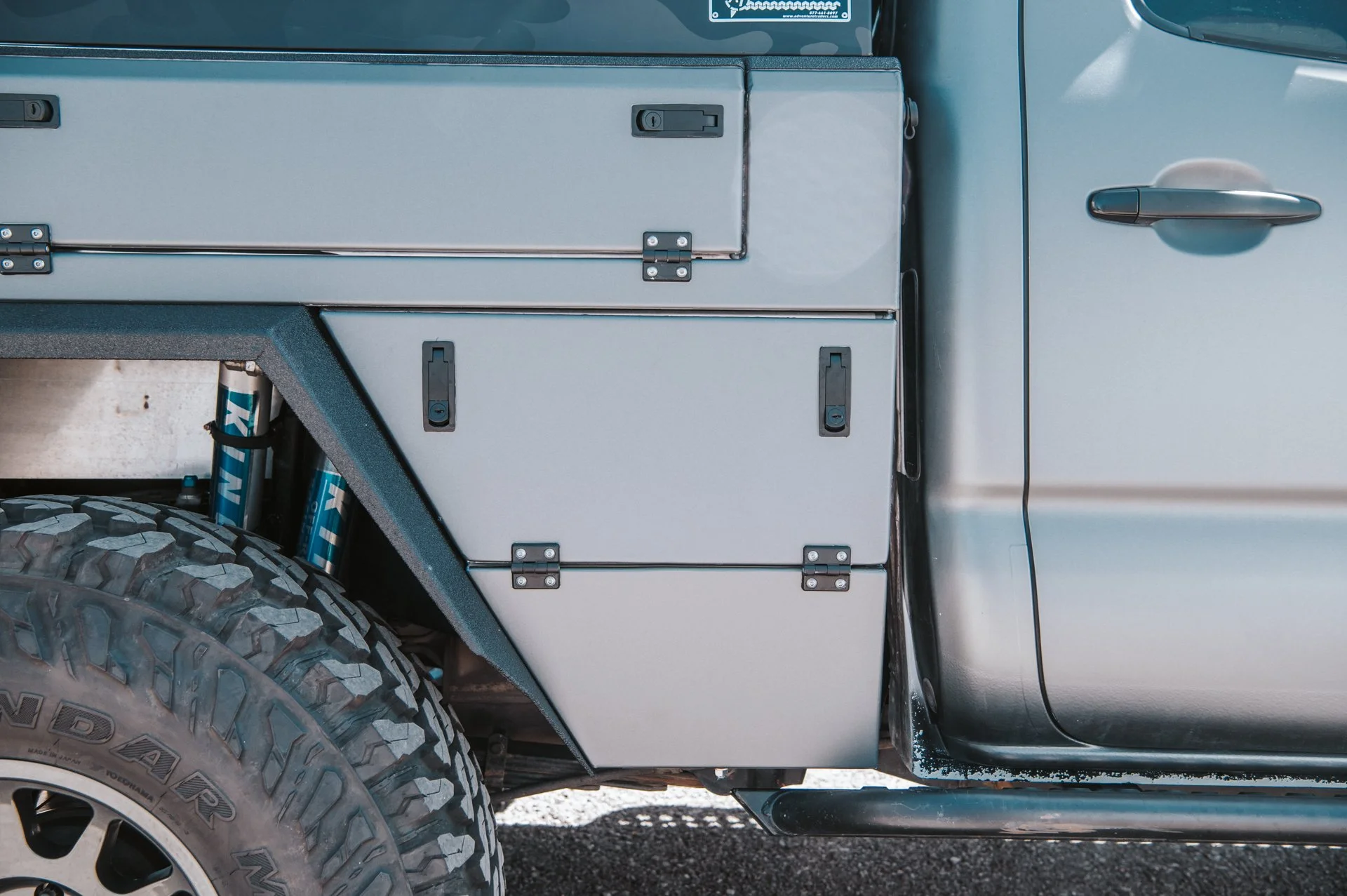 Close-up of the side of a gray utility truck showing storage compartments, a large off-road tire, and part of the vehicle door.