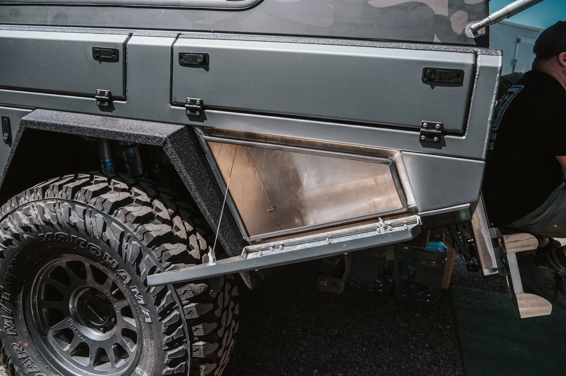 Close-up of a rugged off-road vehicle's side, showing a large tire, a metal storage compartment, and a man sitting next to it, partially visible.