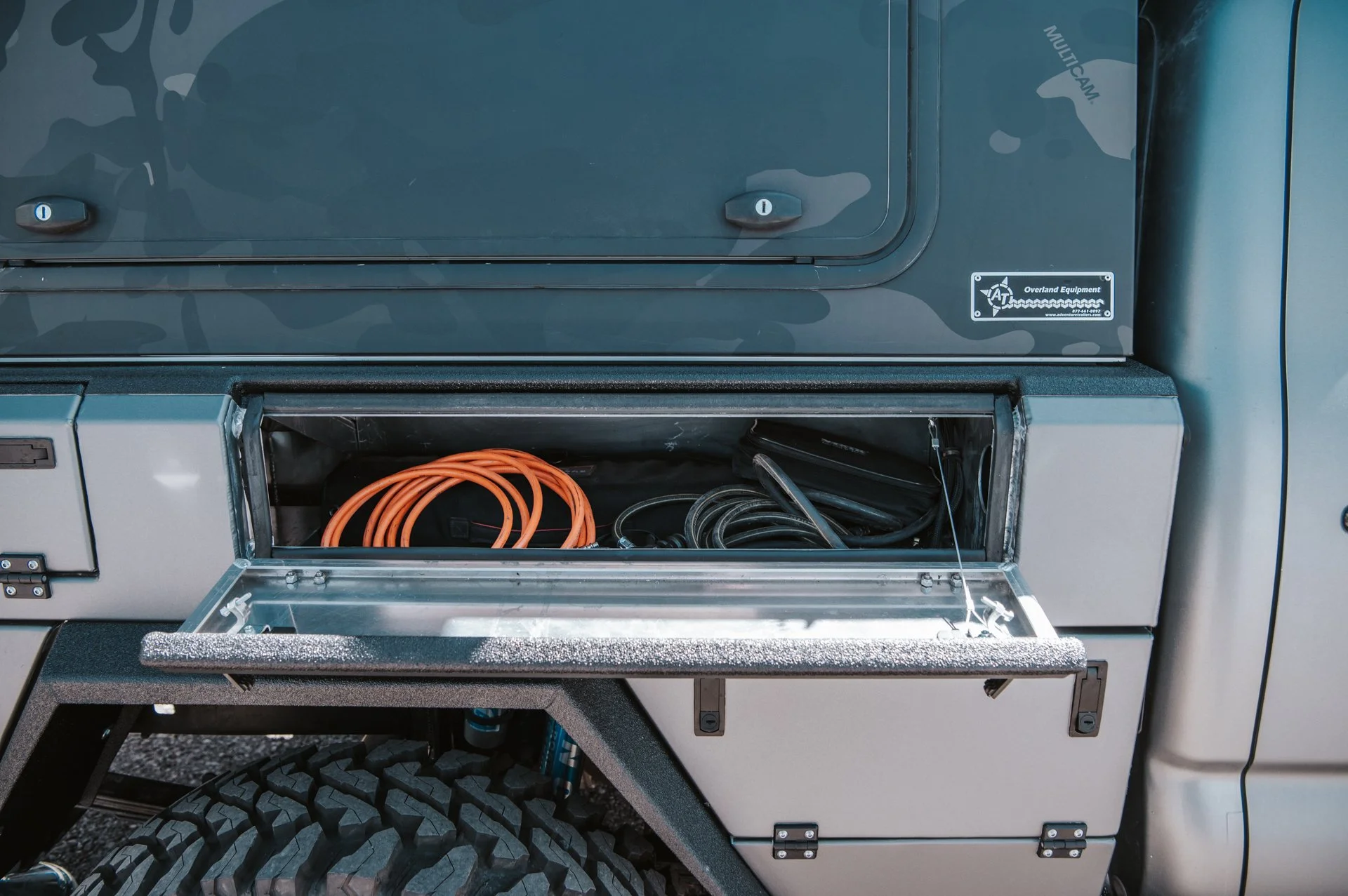 Open storage compartment on the side of a vehicle revealing orange and black cables inside.