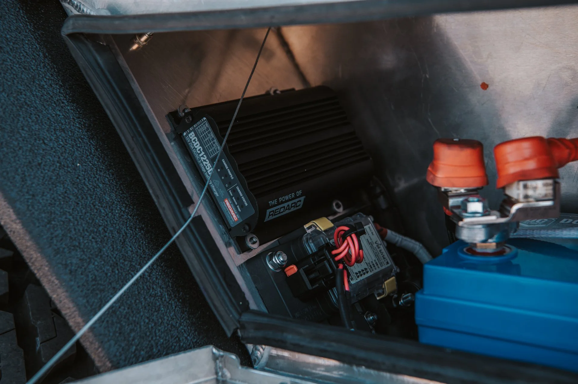 Close-up view of electrical components and wiring inside a vehicle, featuring a black power inverter label, a blue battery box with orange terminal caps, and wiring connections.