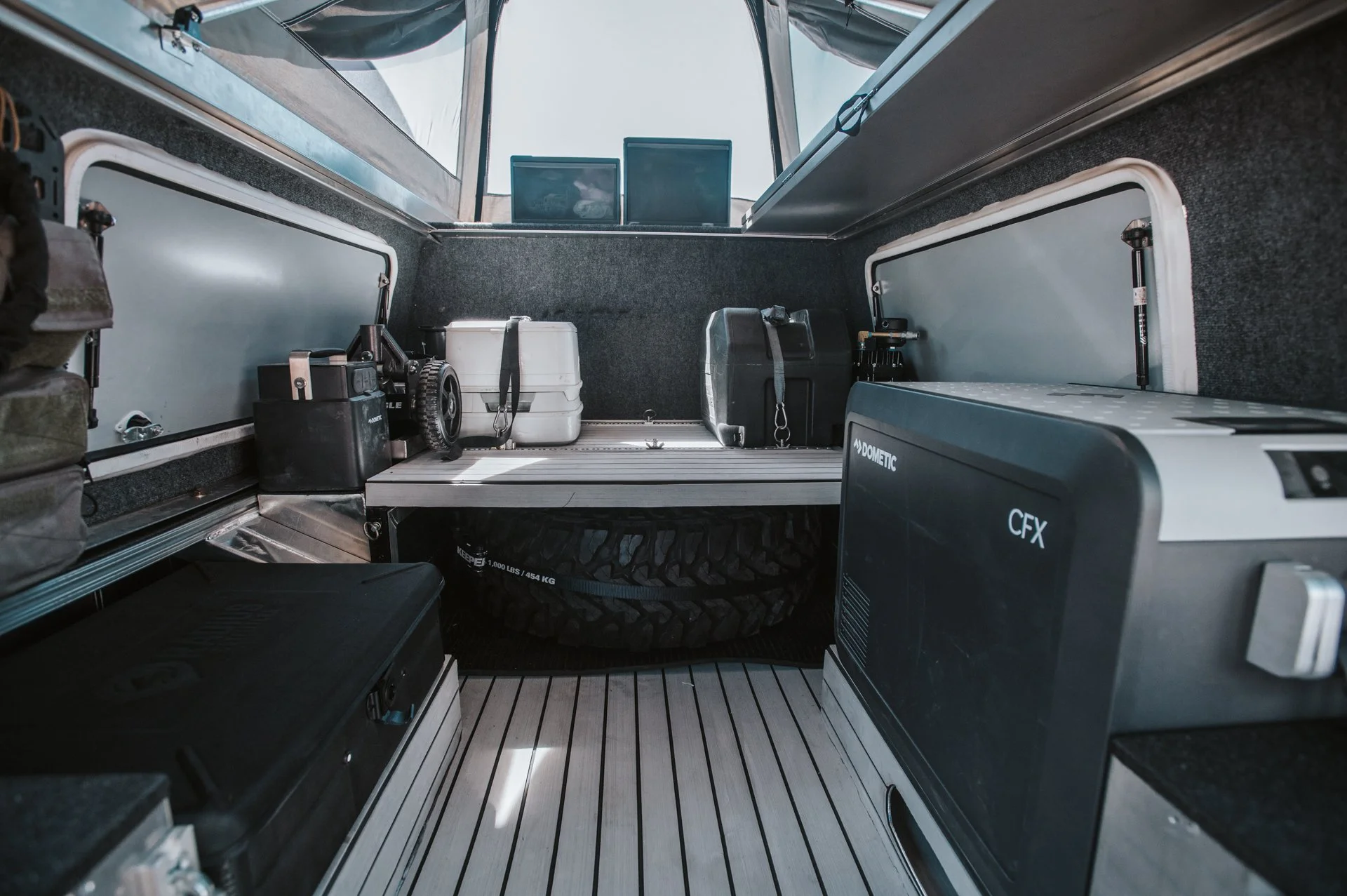 Photo of the interior of a camper van or truck bed storage area, with various equipment and supplies including a tire, a small refrigerator, and containers, with a tent or canopy above.