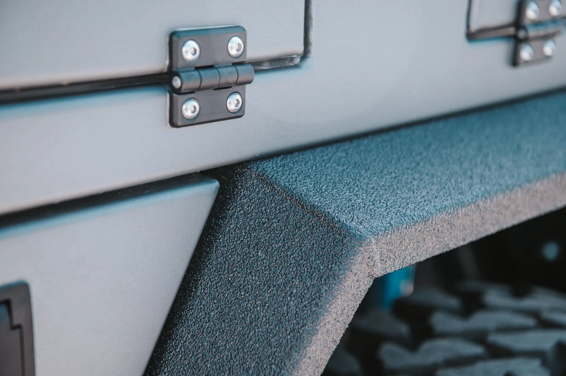 Close-up of a metallic corner and hinge of a gray container or equipment, with textured blue and gray surfaces and a part of a tire visible in the background.
